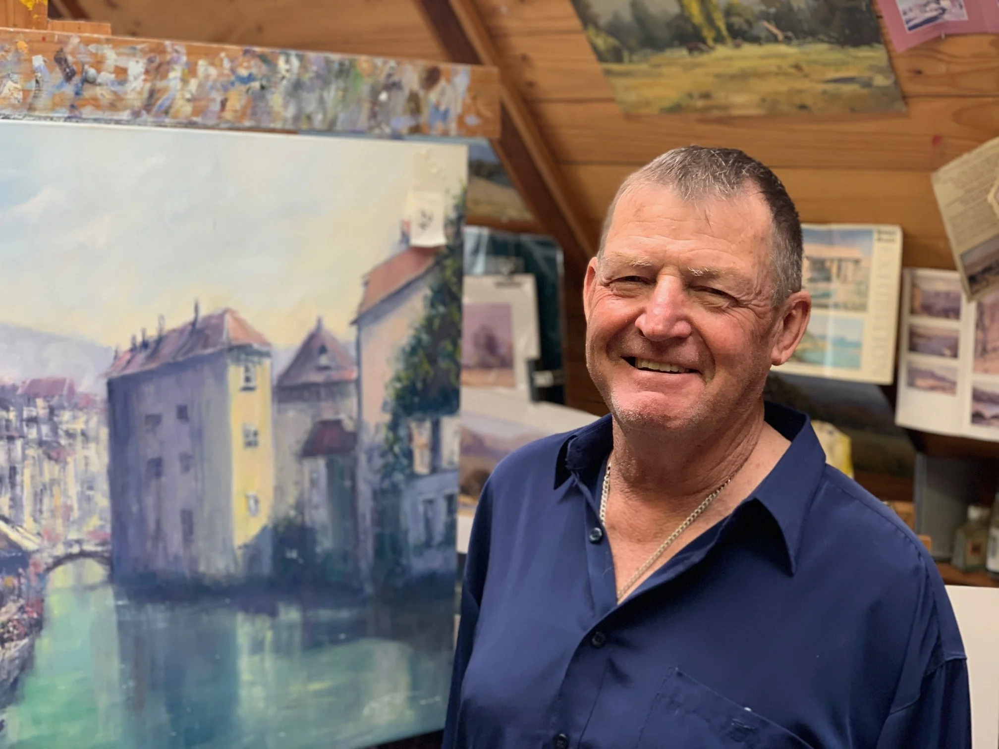 A smiling man with short hair wearing a navy blue shirt, standing in an art studio next to a painting of buildings and a canal.