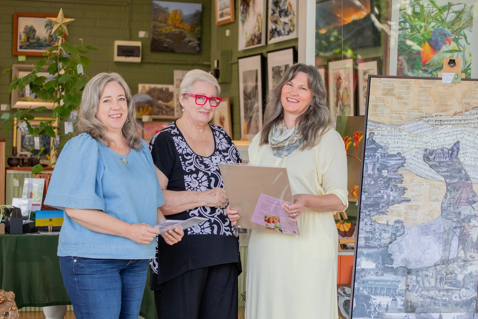Three women standing inside an art gallery, smiling while holding framed pictures. The background shows various paintings and artwork on the wall, with a decorated Christmas tree on the left side.