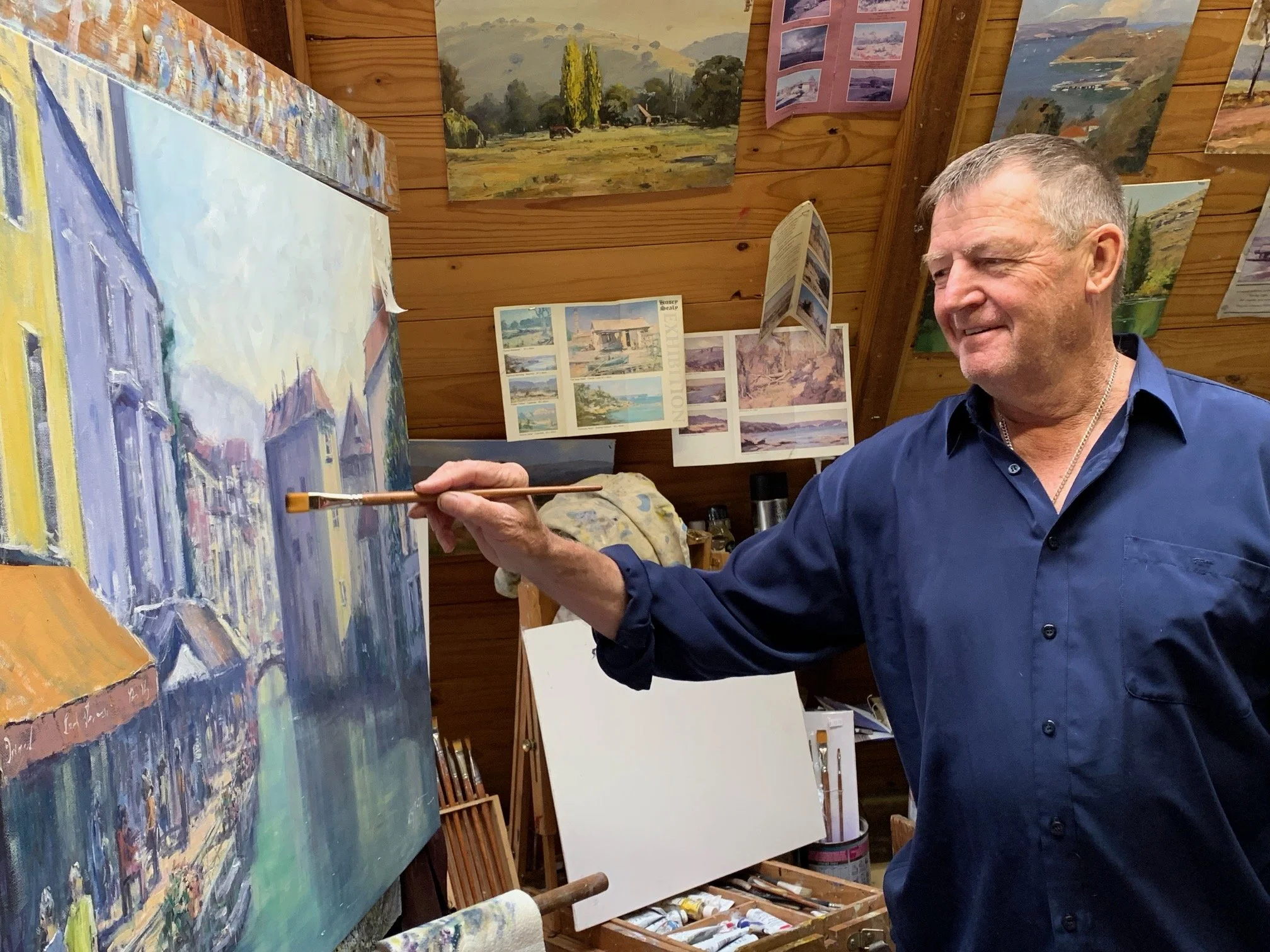 An older man with gray hair painting a landscape on a canvas in an art studio, surrounded by completed artworks and art reference pictures.