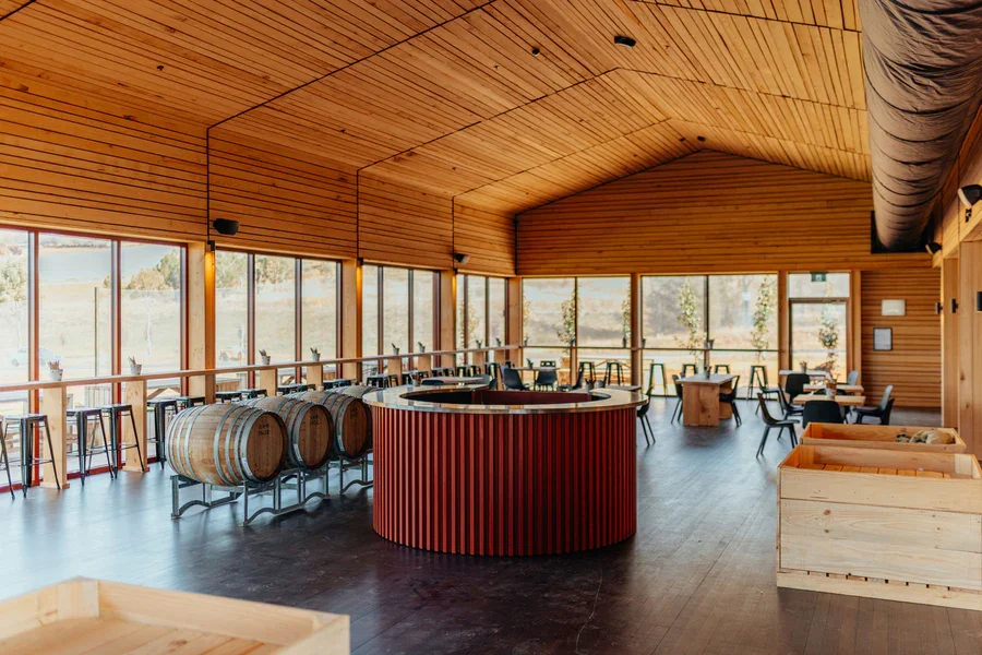 Interior of a cider house with wooden walls and ceiling, large windows, barrels, and tables with chairs.