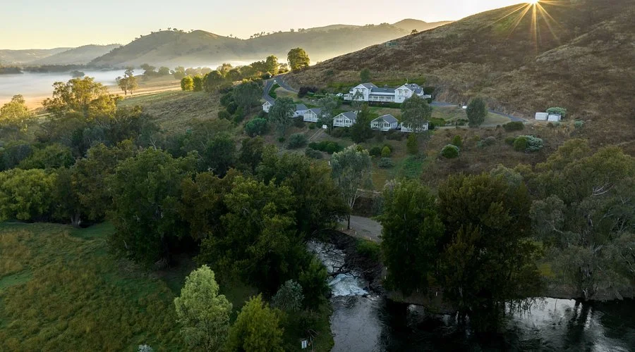 A panoramic view of a valley with lush green fields, scattered trees, and the town of Tumut in the distance, surrounded by rolling hills and mountains under a partly cloudy sky.