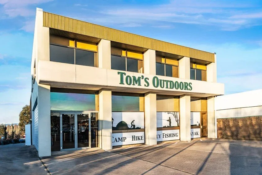 Exterior of a two-story building with signs for outdoor activities like camping, hiking, fishing, kayaking, and hunting, labeled 'Tom's Outdoors'.