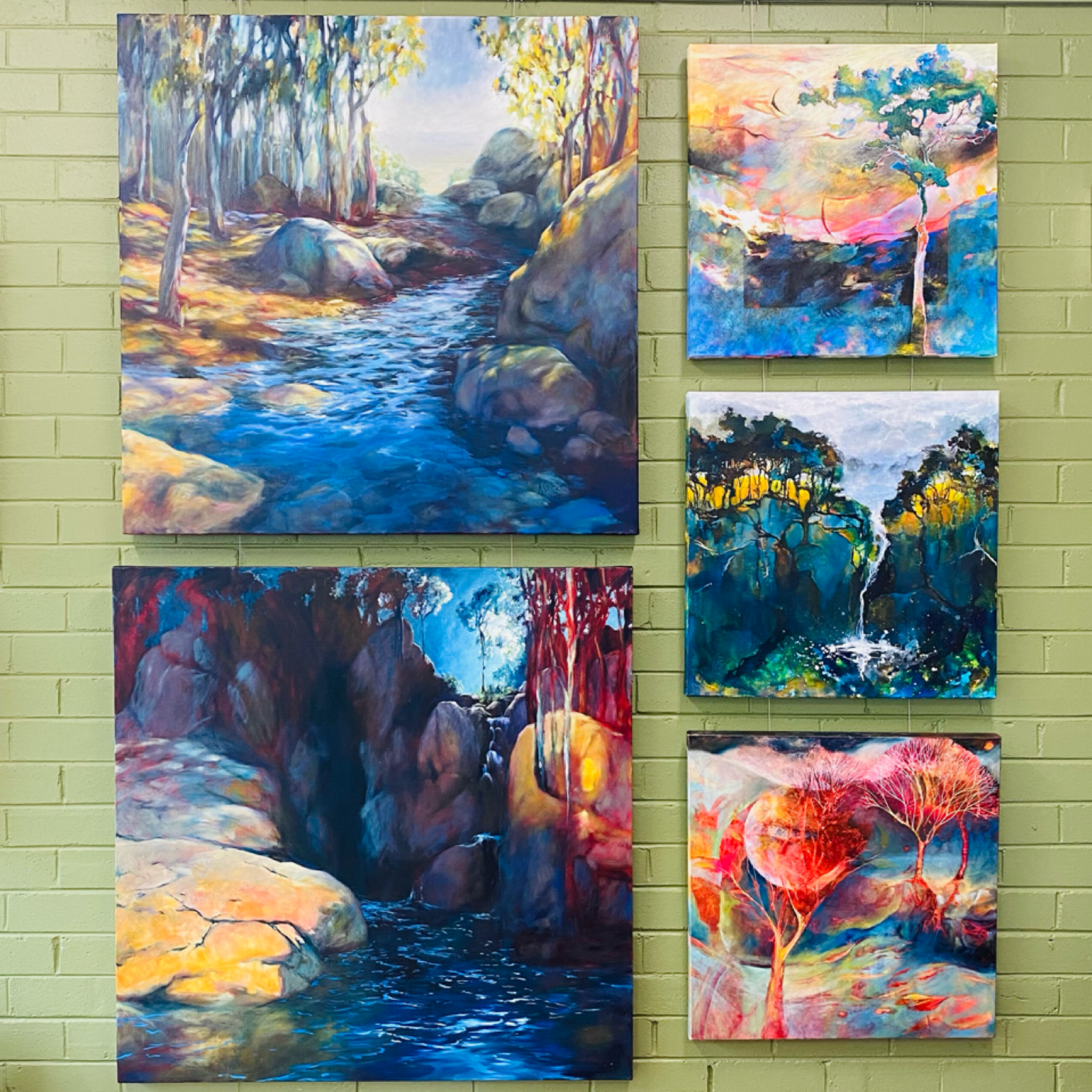 Five colorful landscape paintings depicting rivers, trees, and natural scenery, arranged on a green brick wall.