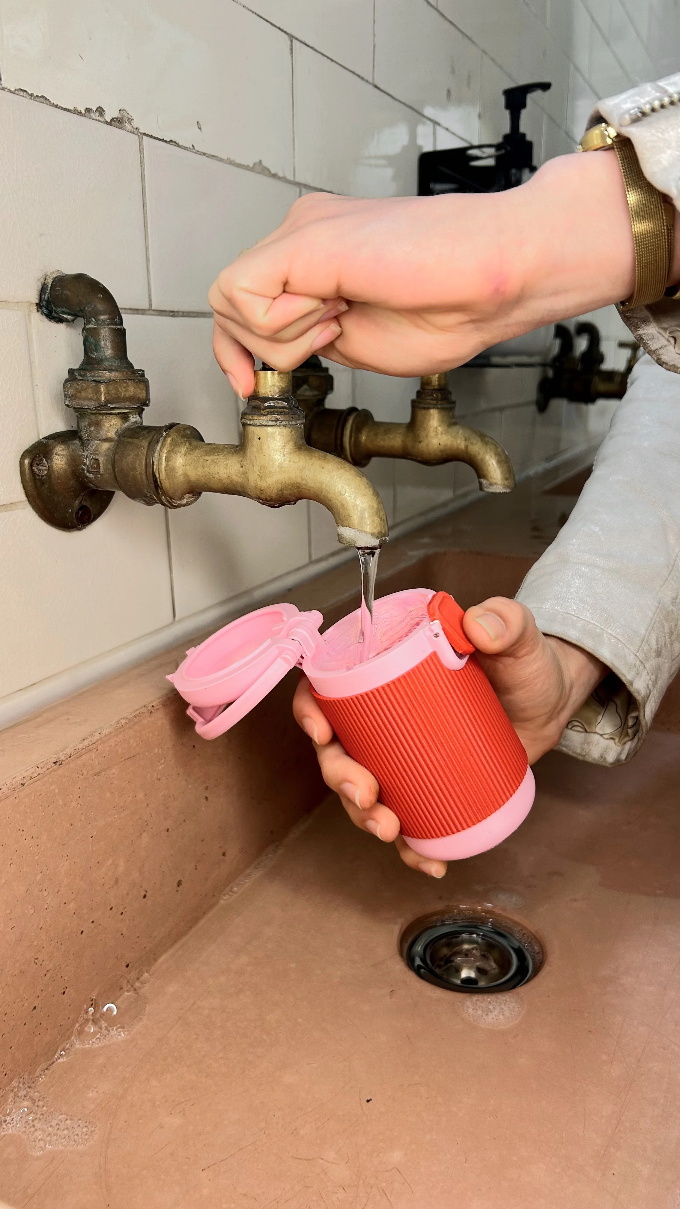 Person filling Halo menstrual cup rinsing pod in the Very Berry colourway under a bathroom tap, with the lid open above a sink