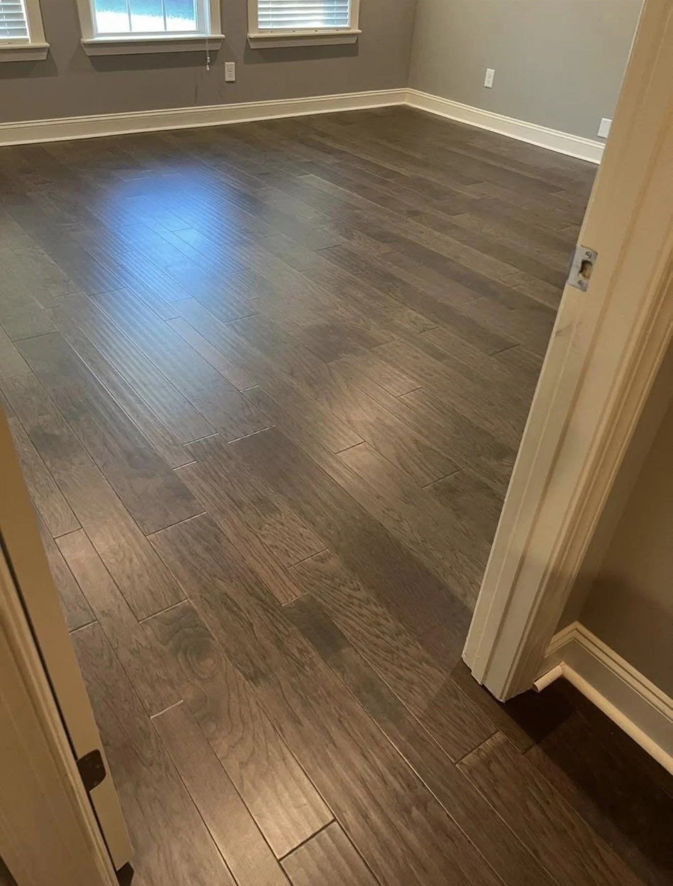 Newly installed dark brown wooden floor in an empty room with gray walls and white baseboards, with two windows on the opposite wall.