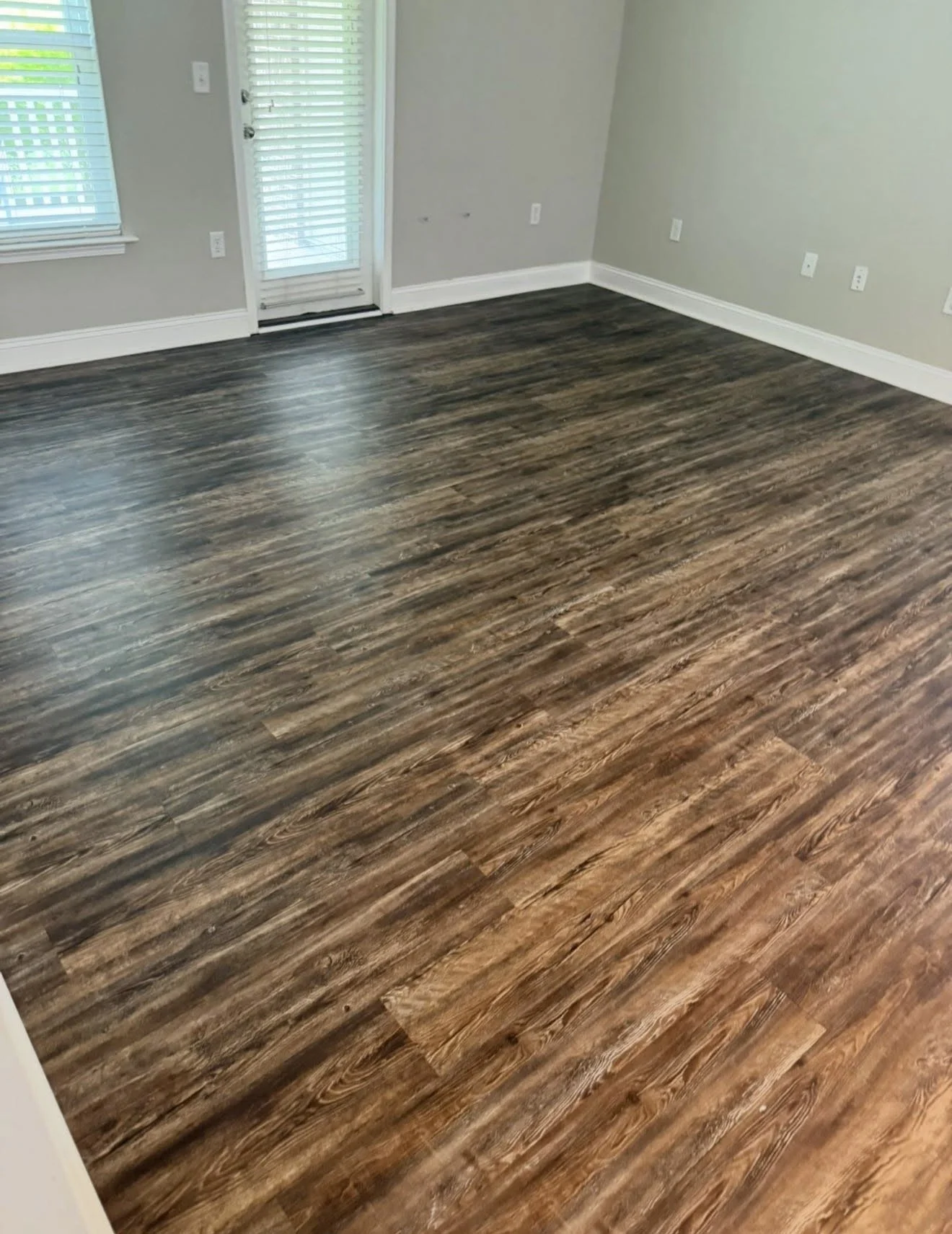 Empty room with dark wood laminate flooring, light gray walls, and white baseboards, with a door and windows with blinds.