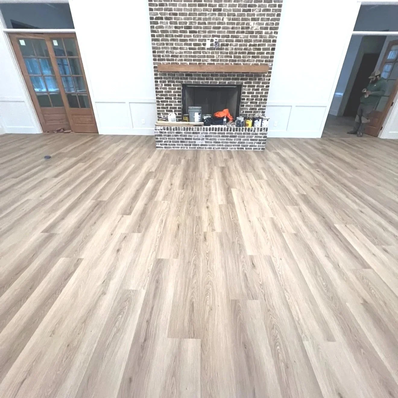 Empty living room with light wooden flooring, brick fireplace, and double wooden doors with glass panels.