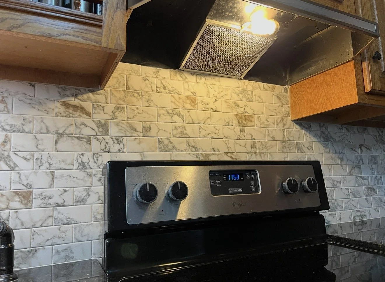 Kitchen stove with control panel and beige marbled tile backsplash, wooden cabinets above, and a black countertop.