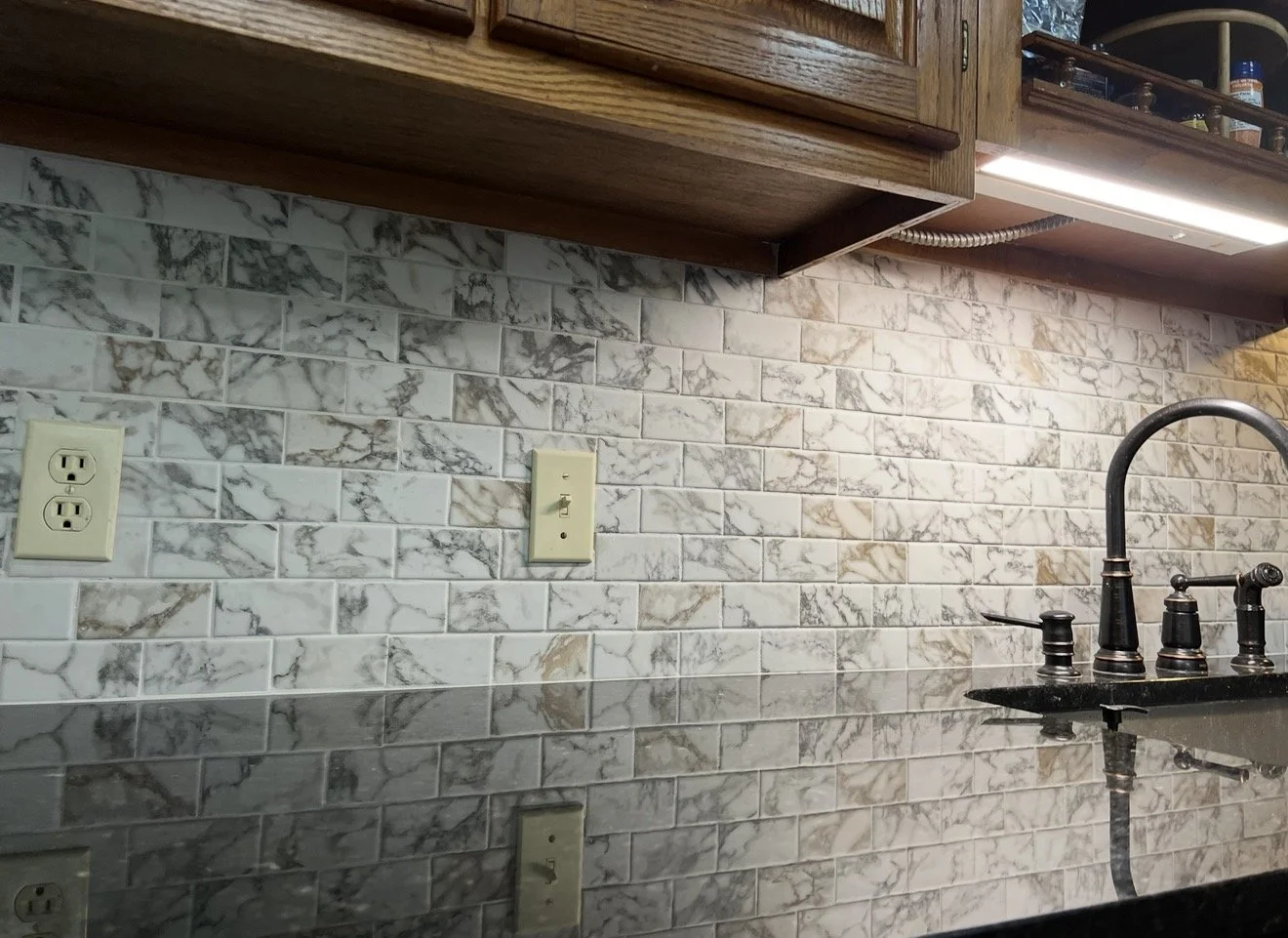 Close-up of a kitchen backsplash with white and gray marble tiles, beneath a wooden cabinet, with electrical outlets and a black kitchen sink faucet visible.