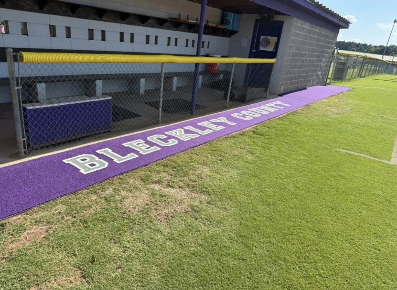 A purple-painted warning area on the ground with the words 'BLACKLEY COUNTY' in white, adjacent to a sports field with green grass, a chain-link fence, and a small building with a door and a purple awning.