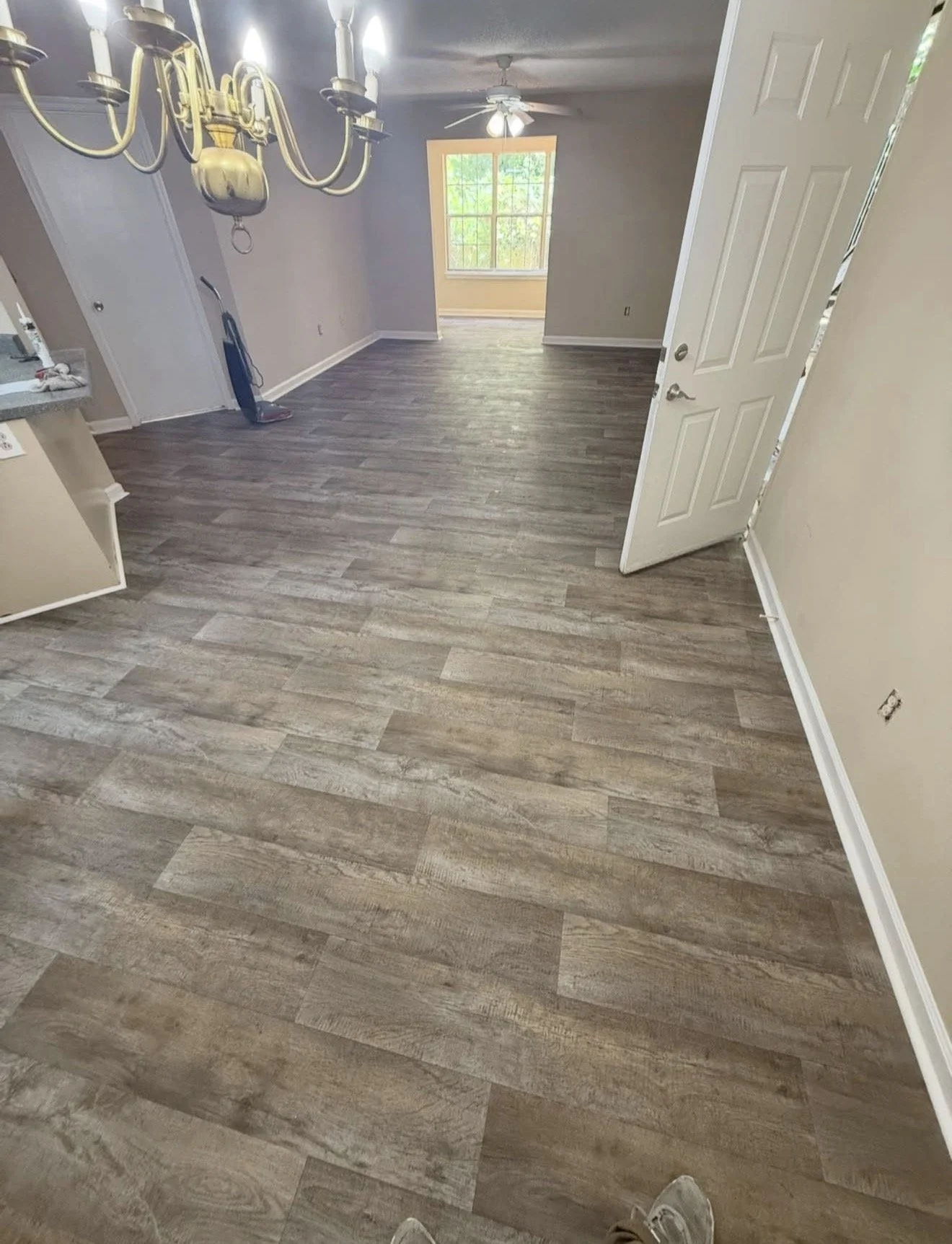 Empty living room with hardwood flooring, an open door on the right, a ceiling fan, large window at the far end, and a chandelier hanging from the ceiling.