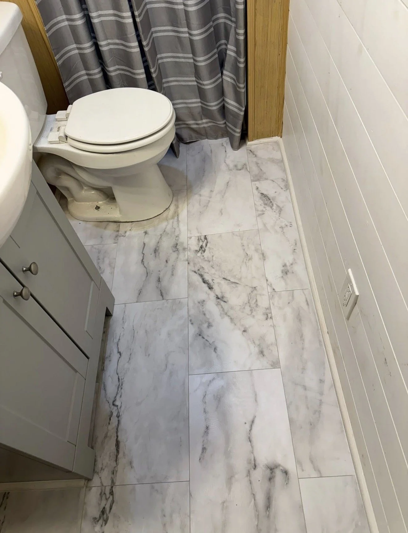 A bathroom corner with a white toilet, a gray cabinet, a gray and white patterned shower curtain, and marble-patterned tile flooring.