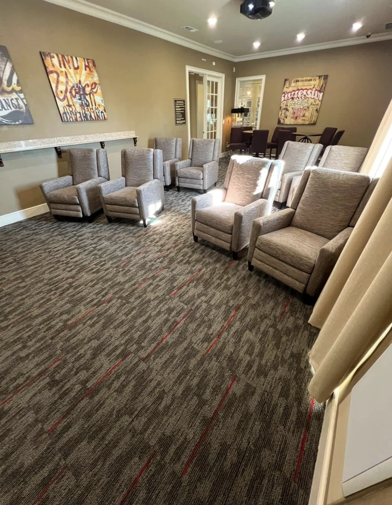 A hotel or conference room with multiple beige armchairs arranged in small groups, a carpeted floor with a pattern, wall art, and a door leading to another room or hallway. Apartment theater