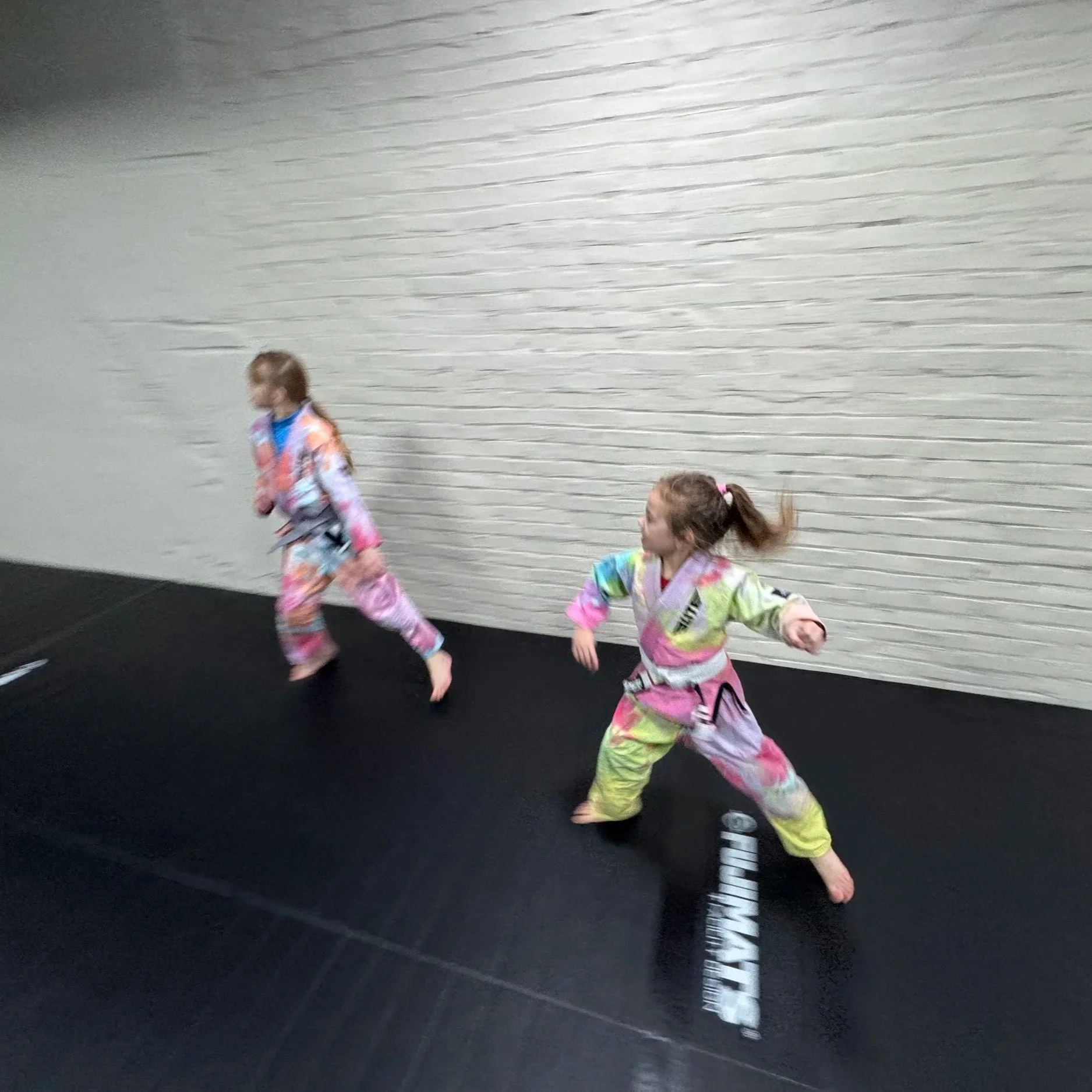 Two young girls practicing martial arts on a black mat indoors, wearing colorful martial arts uniforms.