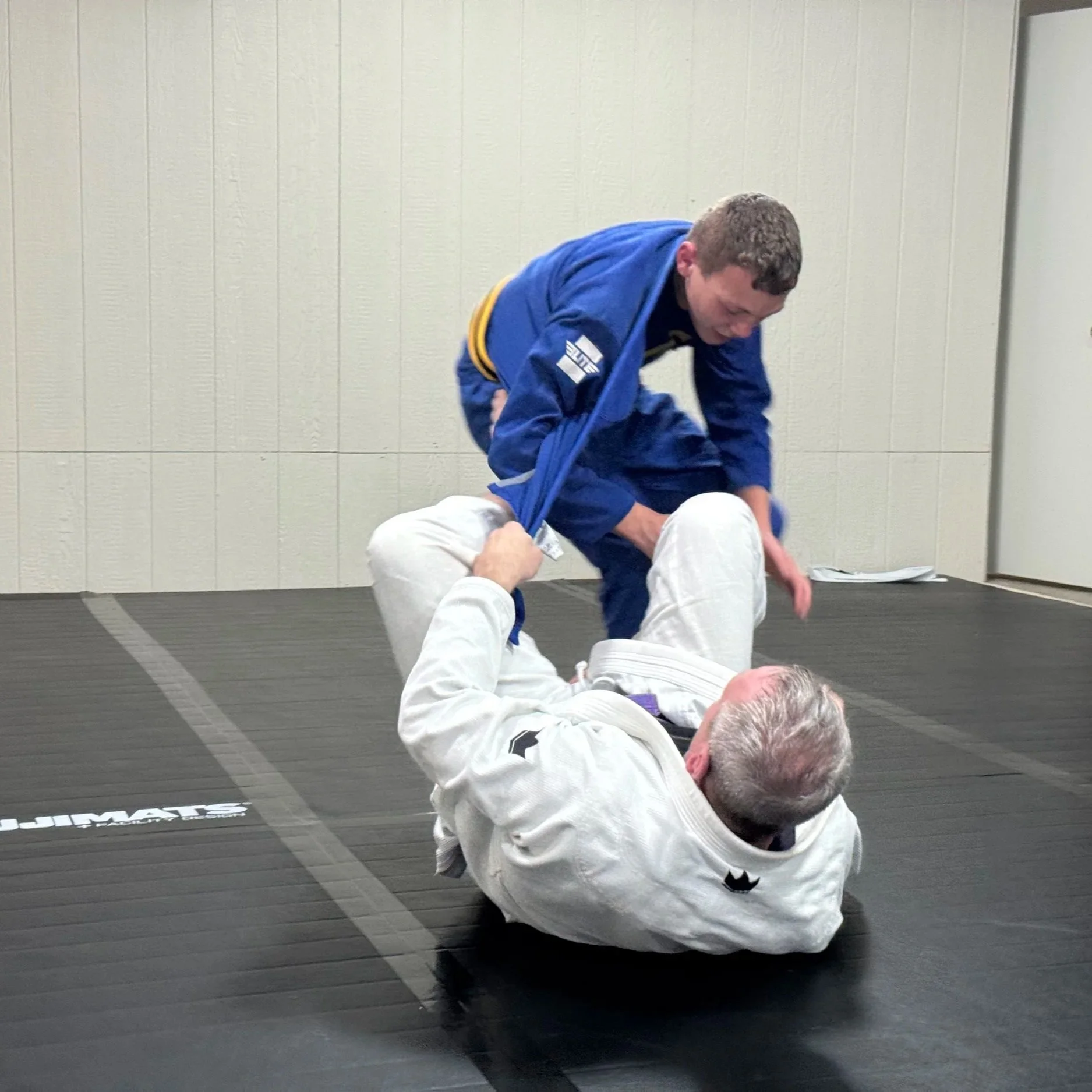 Two men practicing Brazilian Jiu-Jitsu on a mat in an indoor training facility. One man in a white gi is on the ground, while the other man in a blue gi is standing over him.