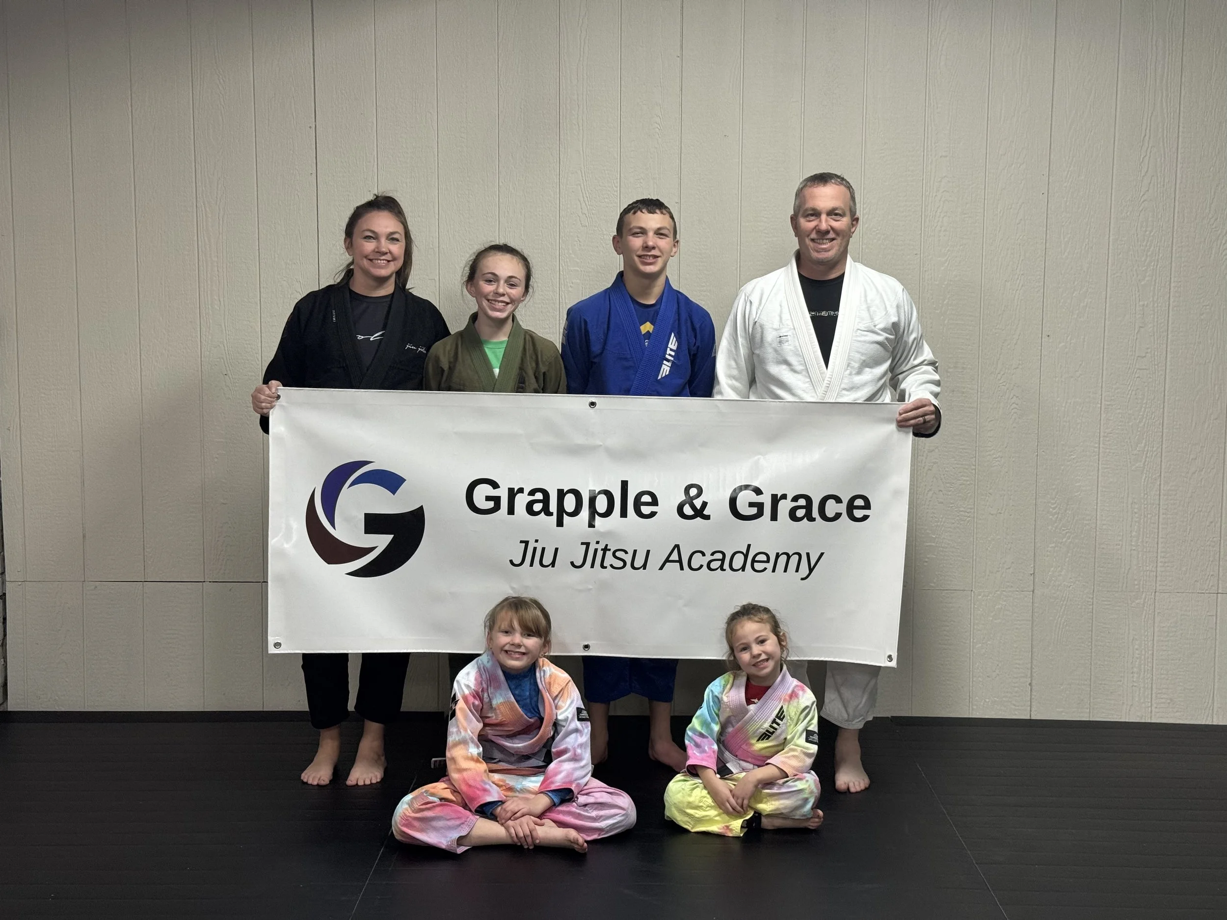 A group of seven people, including children and adults, posing with a banner that reads 'Grapple & Grace Jiu Jitsu Academy.' Five people are standing behind the banner, and two children in colorful gis are sitting on the floor in front. They are all smiling and wearing martial arts gi uniforms, standing against a plain beige wall.