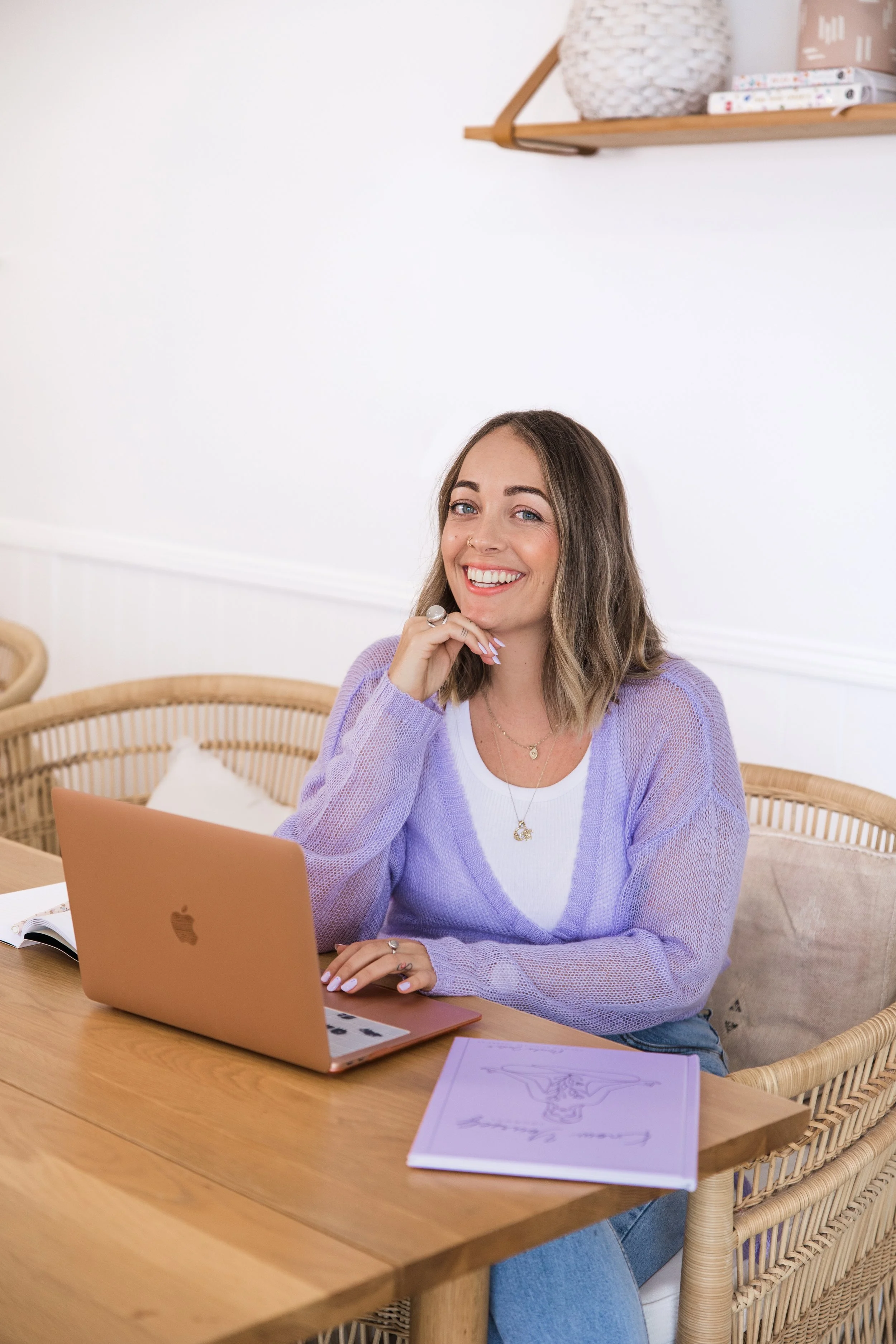 A young woman with long brown hair and blue eyes sitting at a wooden table using a rose gold Apple MacBook. She is smiling and resting her chin on her hand, wearing a lavender sweater, white t-shirt, and layered jewelry, with a classroom or workspace background.