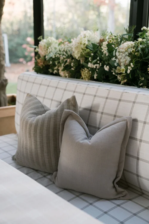 A cozy seating area with two beige pillows on a plaid upholstered bench, above which is a window with a flower box filled with white and green flowers.
