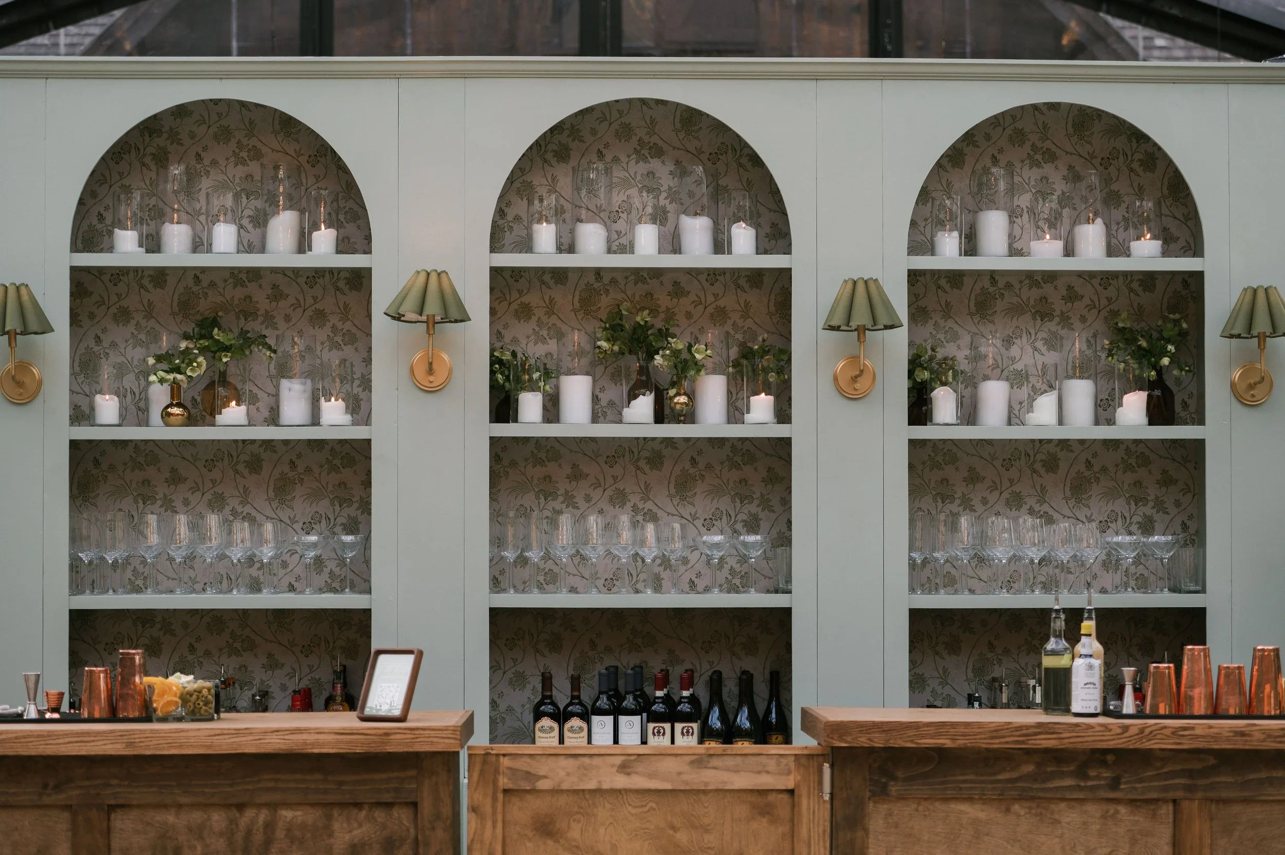 A bar with a light green shelving unit decorated with white candles and greenery, patterned wallpaper inside the shelves, and bottles of wine on the counter below.