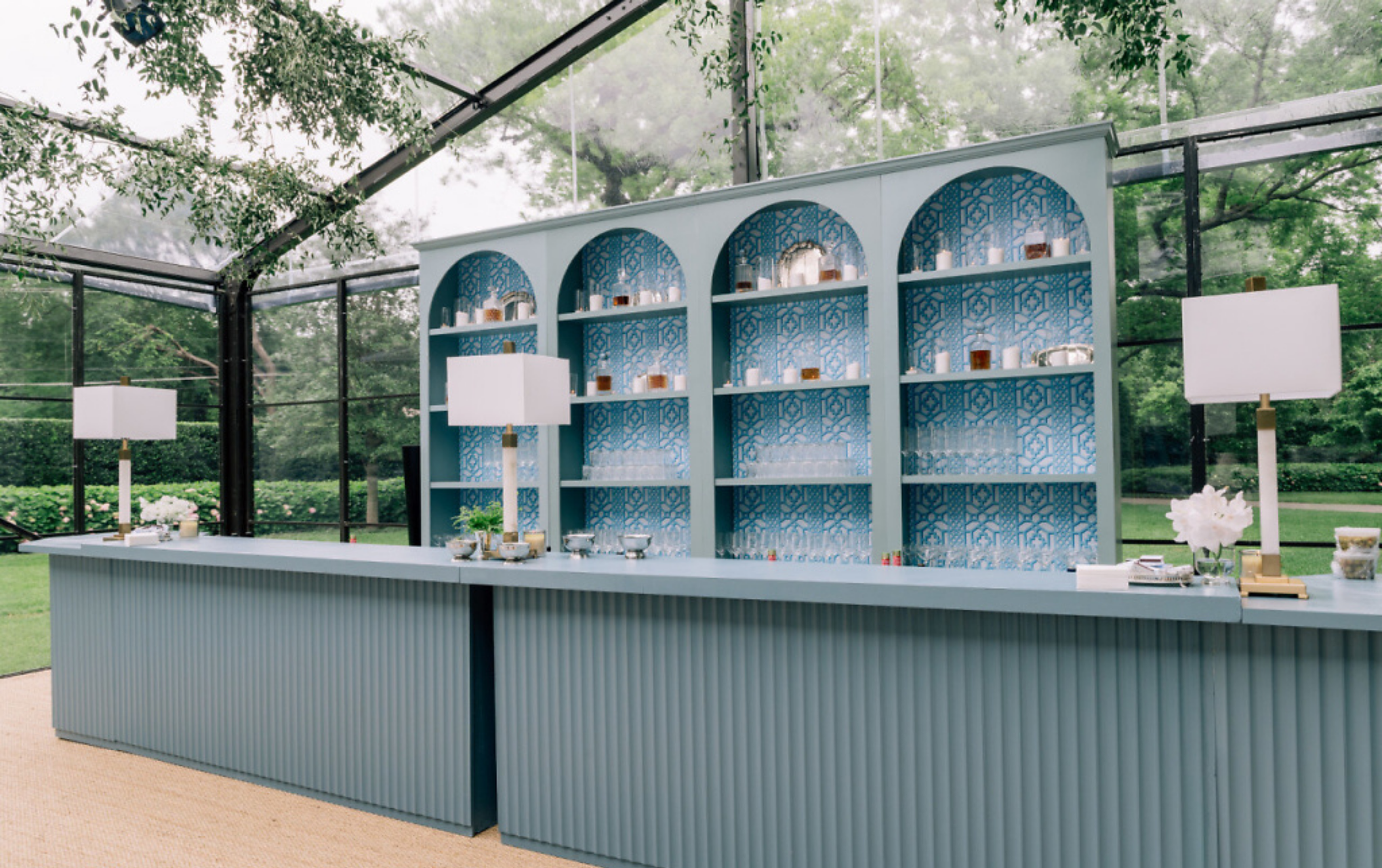 A blue and white bar with decorative geometric cutouts, set inside a glass conservatory with trees visible outside, featuring pendant lamps and assorted bottles and glasses on the shelves.
