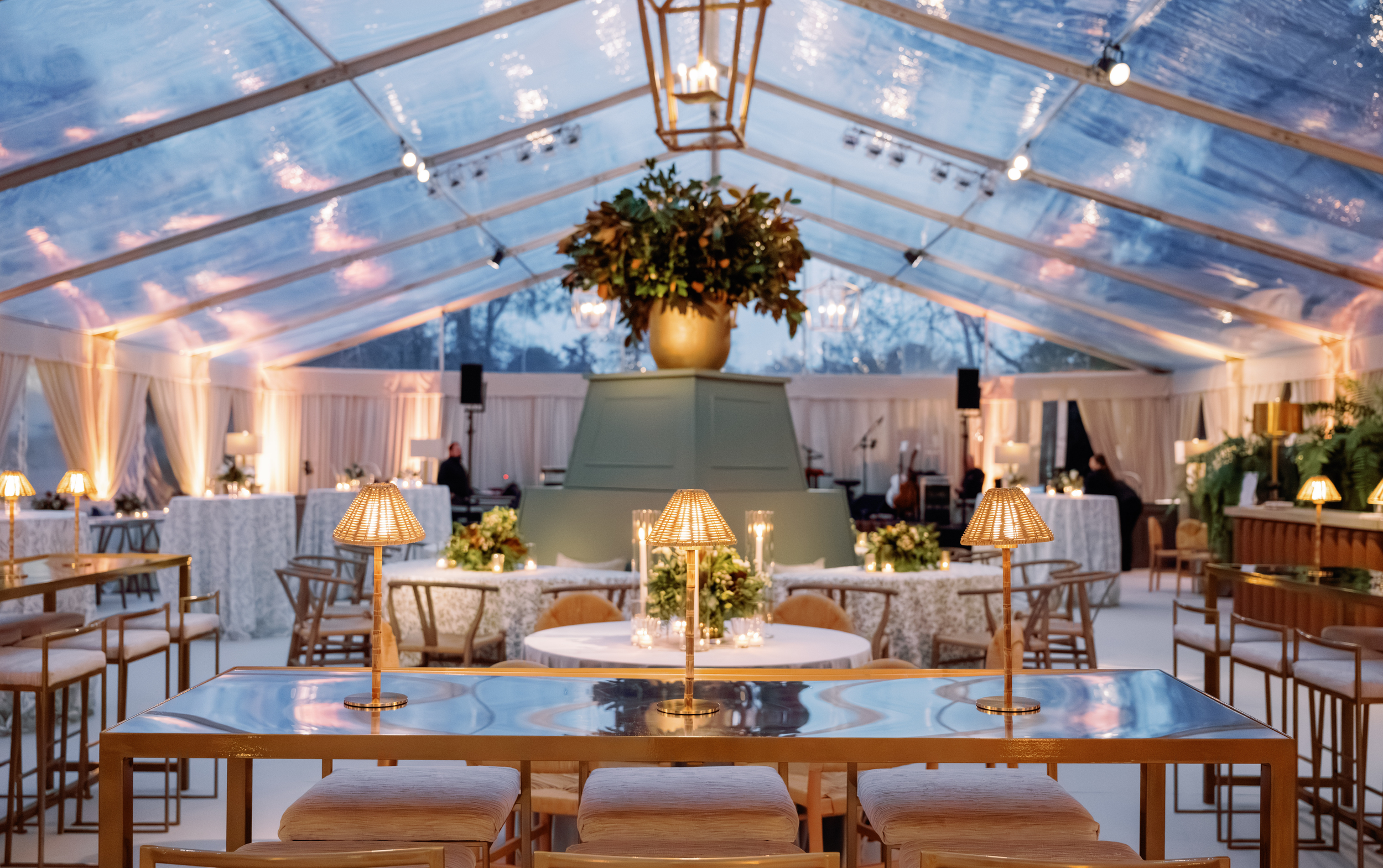 Interior view of a decorated event tent with a high transparent ceiling, string lights, a large floral arrangement in a gold vase, round tables with white tablecloths, candles, and seating for guests.