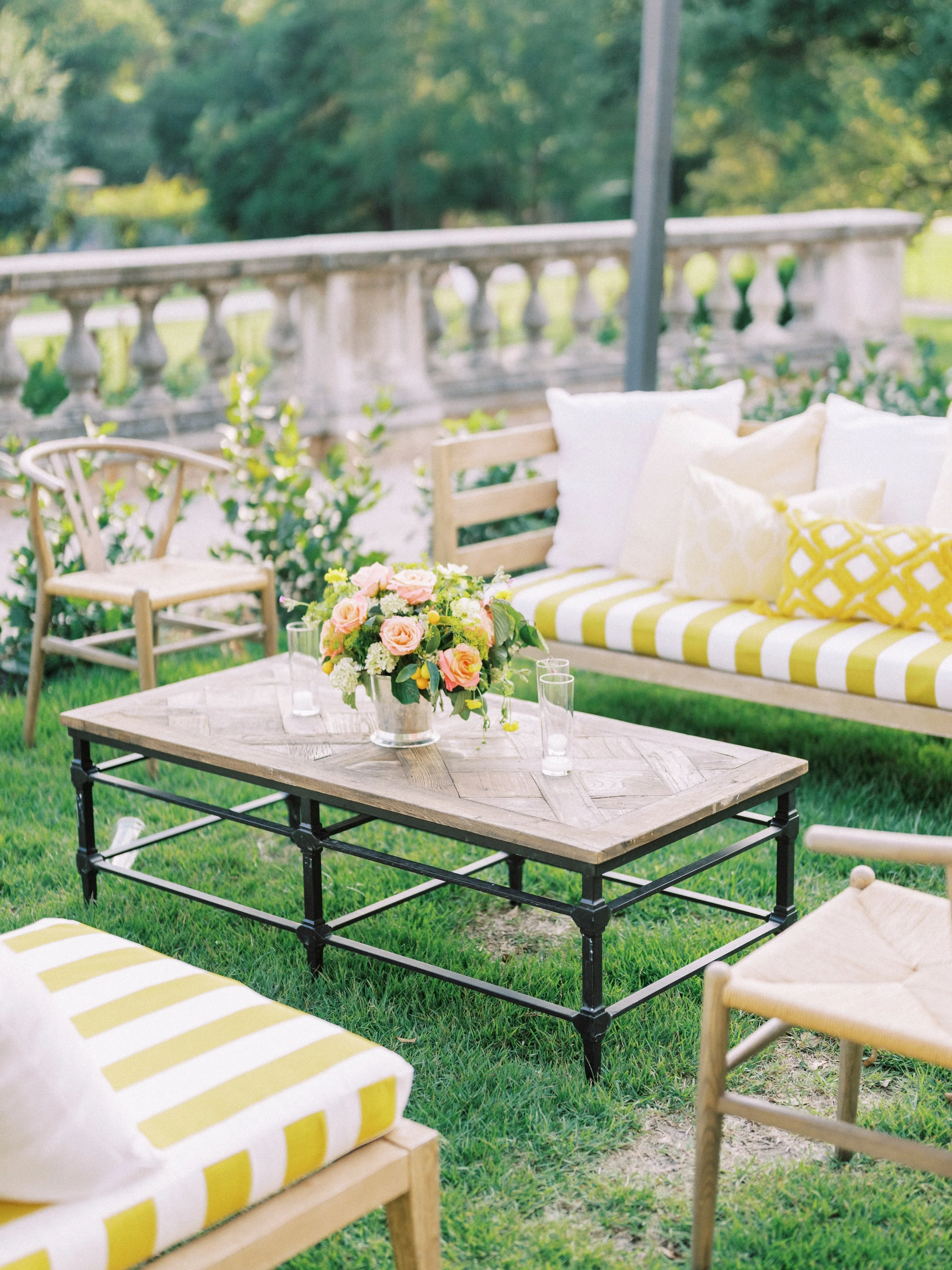 Outdoor patio setup with a wooden coffee table, a bouquet of pink roses, and seating with yellow and white striped cushions surrounded by grass and trees.