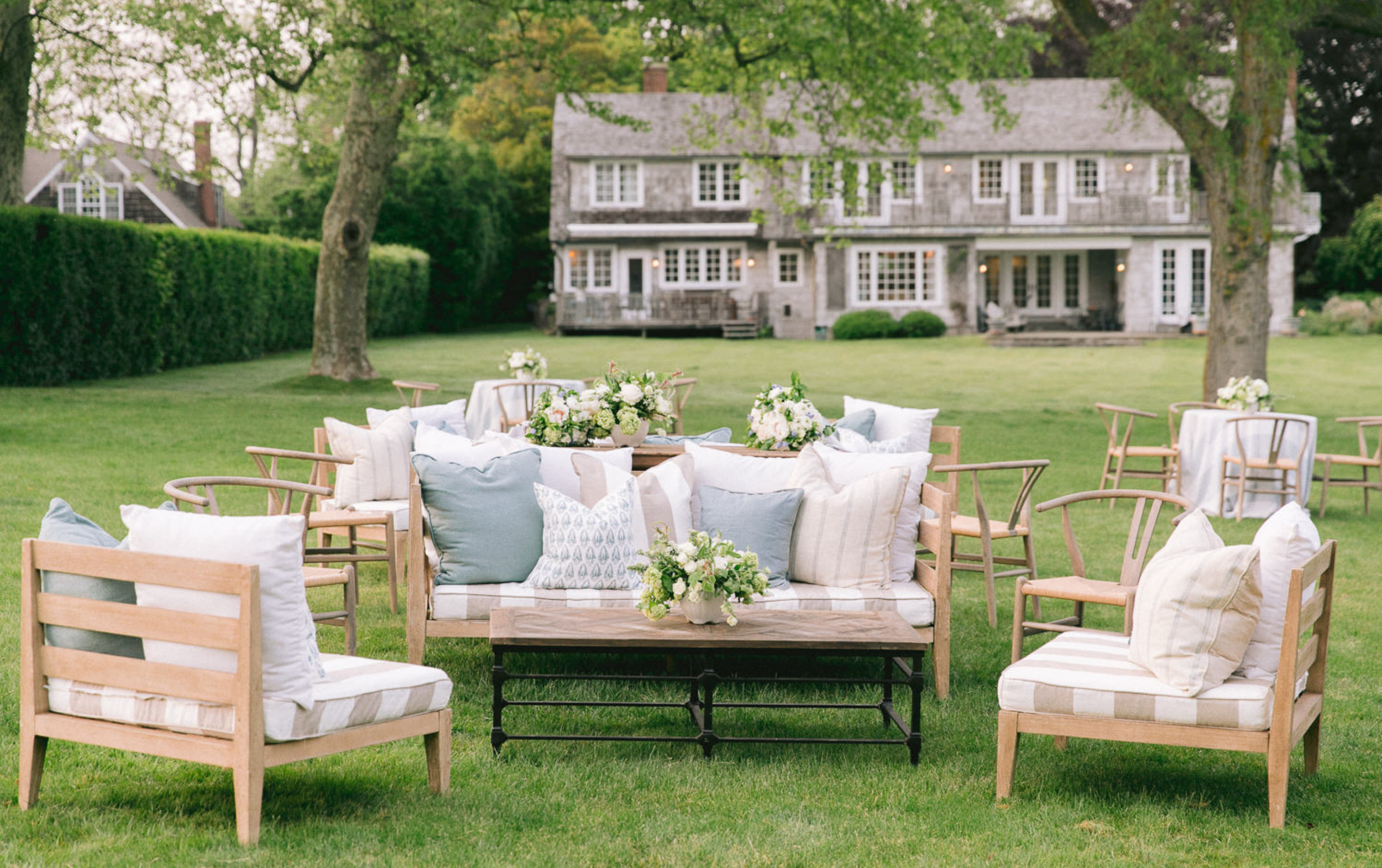 Outdoor garden setting with arranged wooden chairs and couches decorated with pillows, along with flower arrangements on tables, in front of a large house with a rustic appearance surrounded by trees and lush greenery.