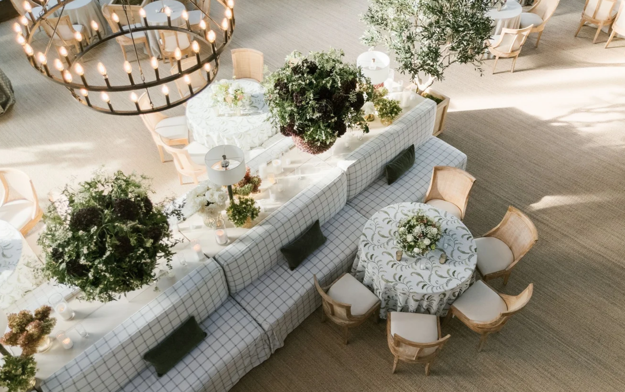 An elegantly decorated banquet hall with round tables covered in white and patterned tablecloths, surrounded by wooden chairs with white cushions. The tables have floral centerpieces, candles, and glassware. A large floral arrangement hangs down over