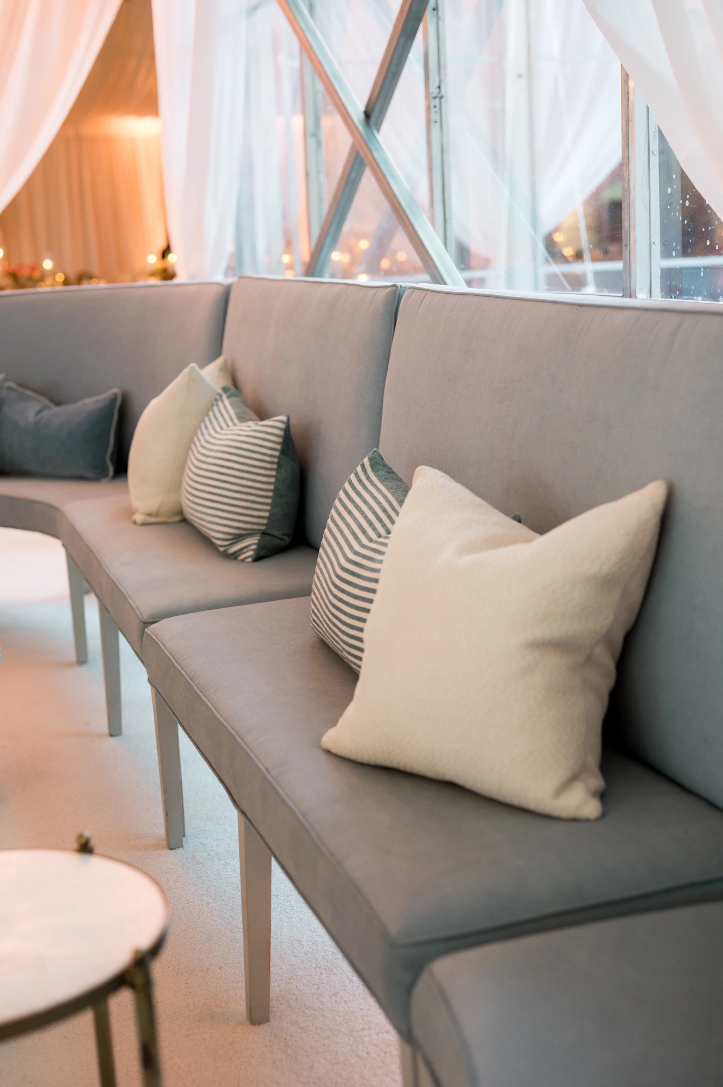 A modern lounge area with a long gray upholstered sofa adorned with seven decorative pillows in various shades of gray, beige, and striped patterns. The sofa is positioned in front of large windows with sheer white curtains, allowing soft natural light to fill the room. Partially visible in the foreground is a round side table with a decorative item on top.