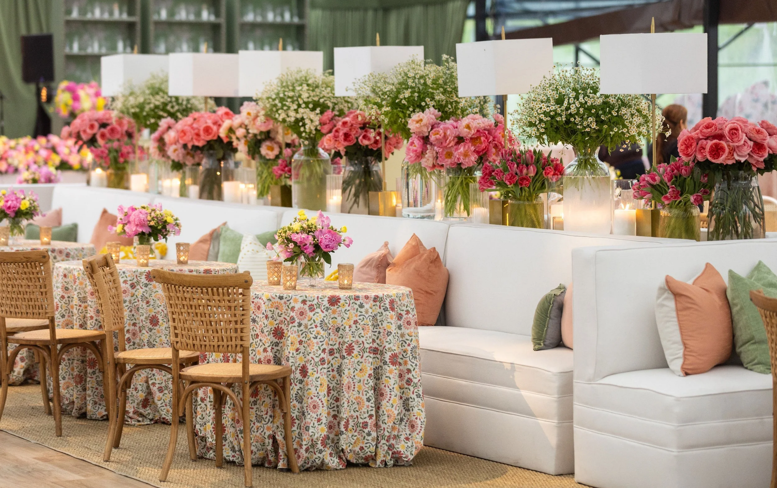 A decorated event space with large floral arrangements in vases, candles, tables with floral tablecloths, and chairs with pillows.