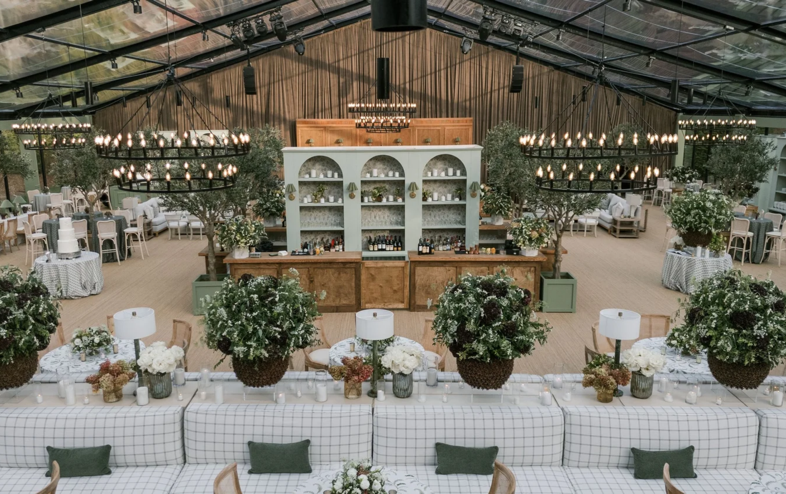 Elegant indoor event space with floral arrangements, candle decorations, and a bar area, featuring high ceilings with exposed beams and chandeliers.