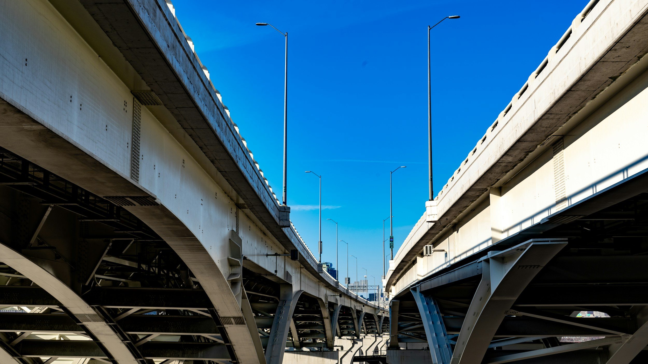 Underbridge view of highways with multiple lanes, overhead streetlights, and a clear blue sky in an urban setting.
