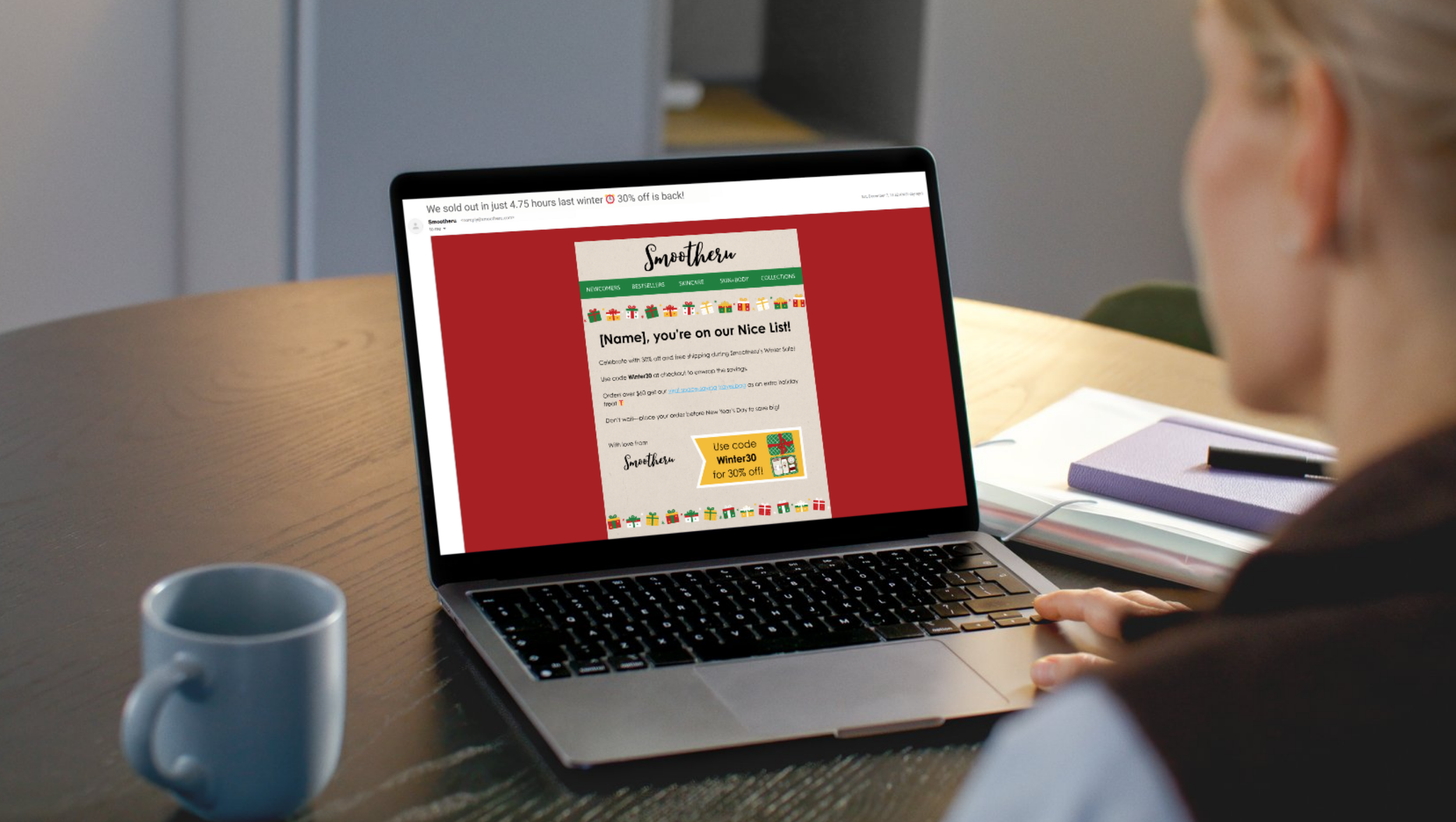 Person viewing a promotional email from Smotheeru on a laptop on a wooden table, with a white mug, a purple notebook, and a pen nearby.