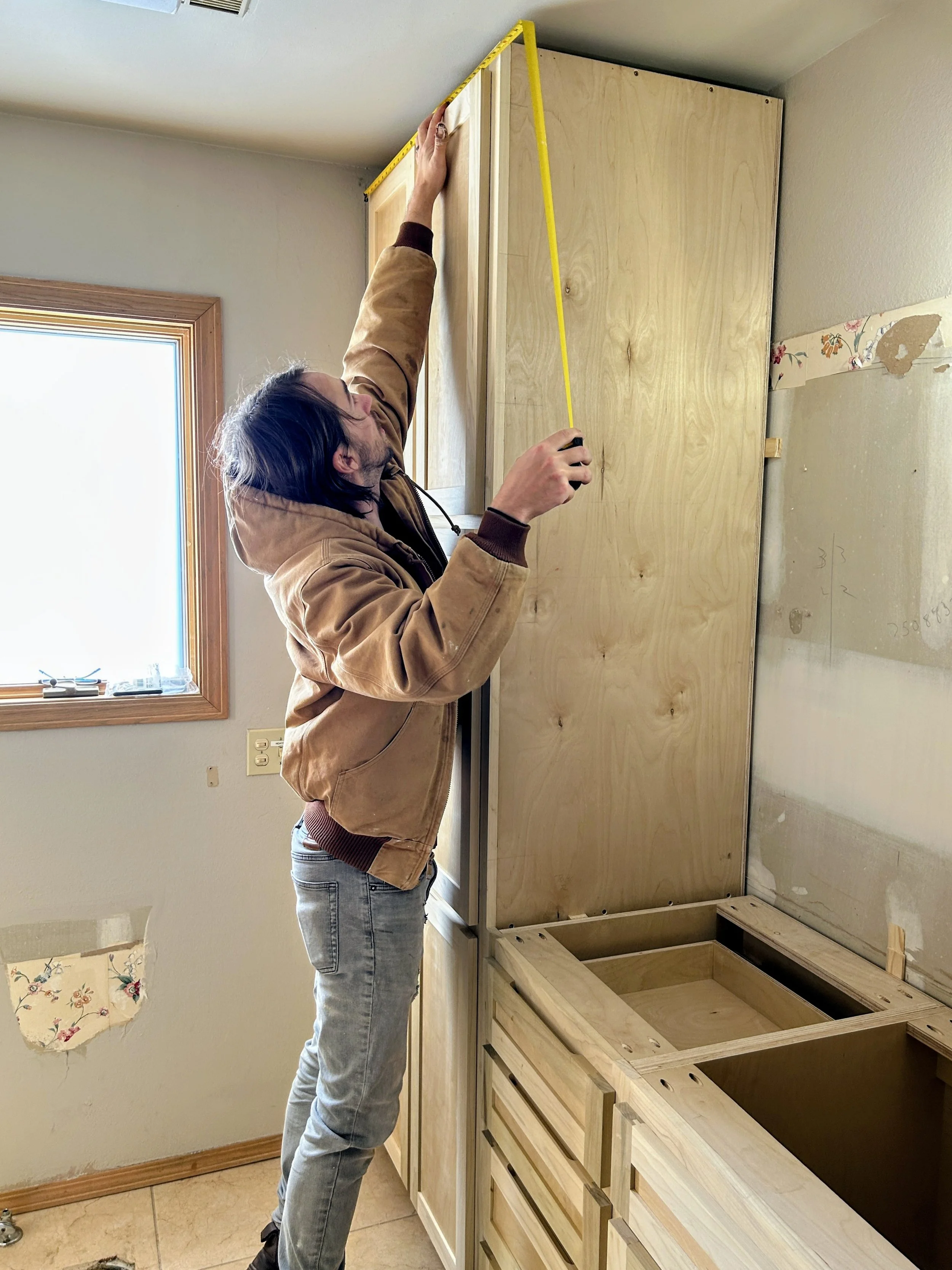 A man measuring the height of a tall wooden cabinet in a kitchen under construction or renovation.