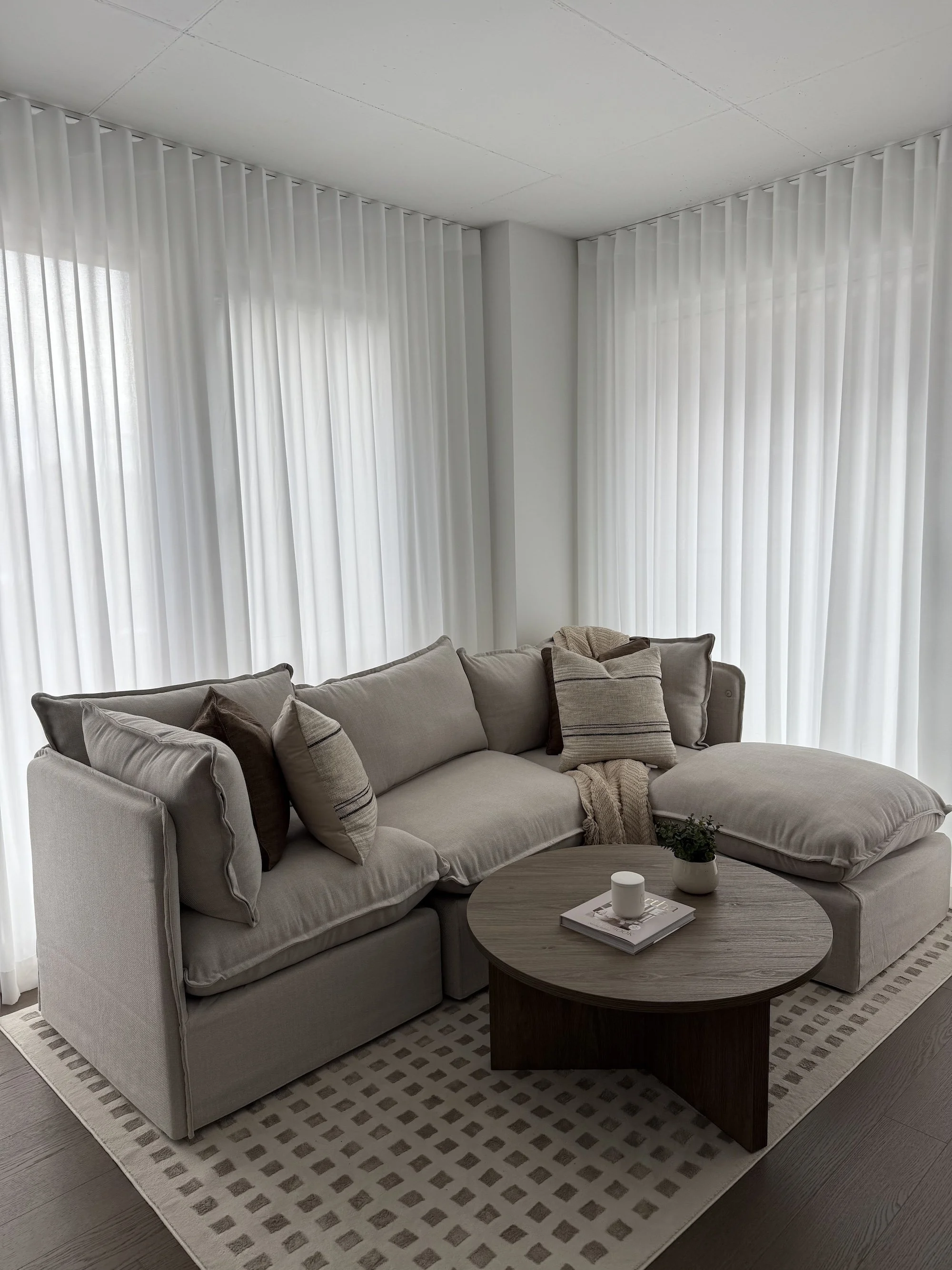 Living room with beige sectional sofa, decorative pillows, a throw blanket, round wooden coffee table with a plant, candle, and magazine, light-colored curtains, and a textured area rug.