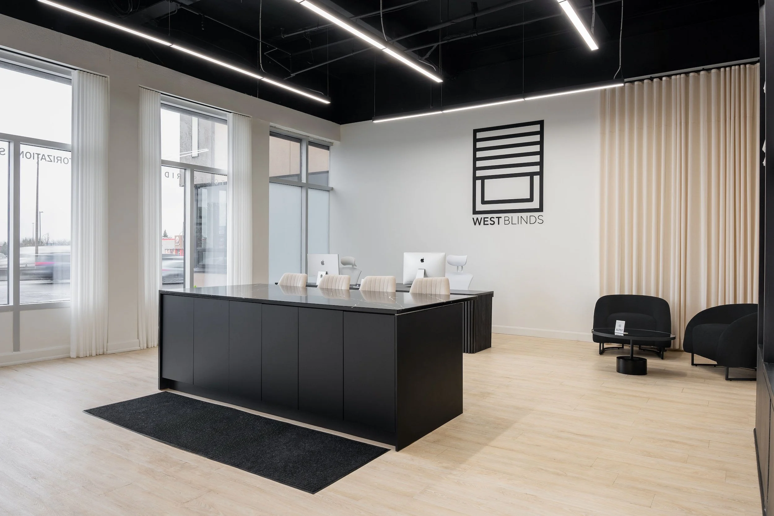Modern reception area with large windows, white walls, and black ceiling, featuring a sleek black reception desk with four white chairs and two black lounge chairs, a logo on the wall reading 'WEST BLINDS,' and beige curtains on the right side.