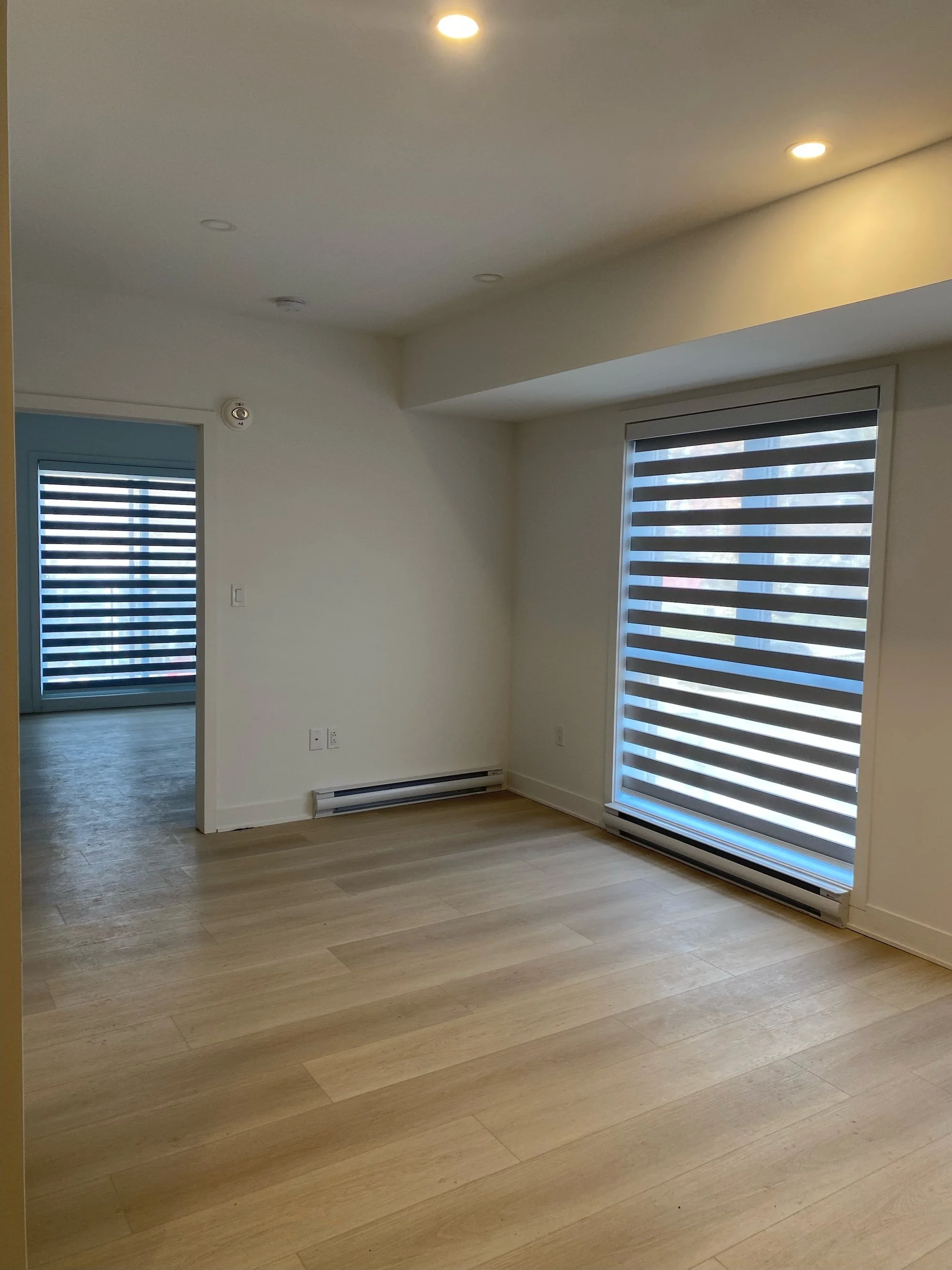 Empty room with light wood flooring, white walls, large window with horizontal blinds, and a doorway leading to another room with a similar window and blinds.