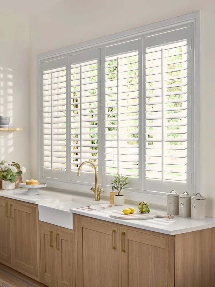 Kitchen window with white plantation shutters and a white farmhouse sink, wooden cabinets, and a gold faucet.
