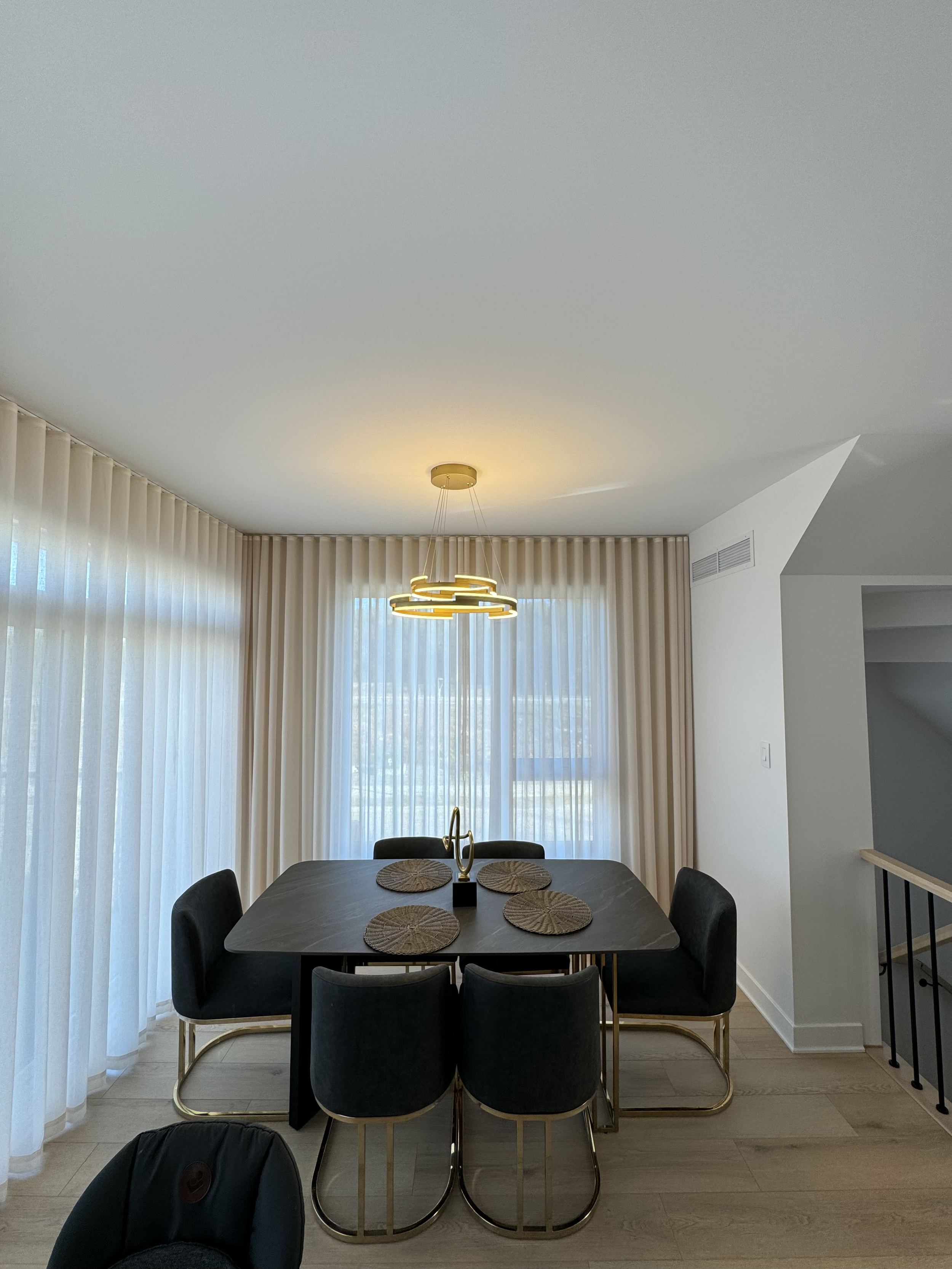 Dining room with a black rectangular table, six black chairs with gold frames, and six woven placemats. A modern gold chandelier hangs above, and there are large sheer curtains covering the windows in the background.