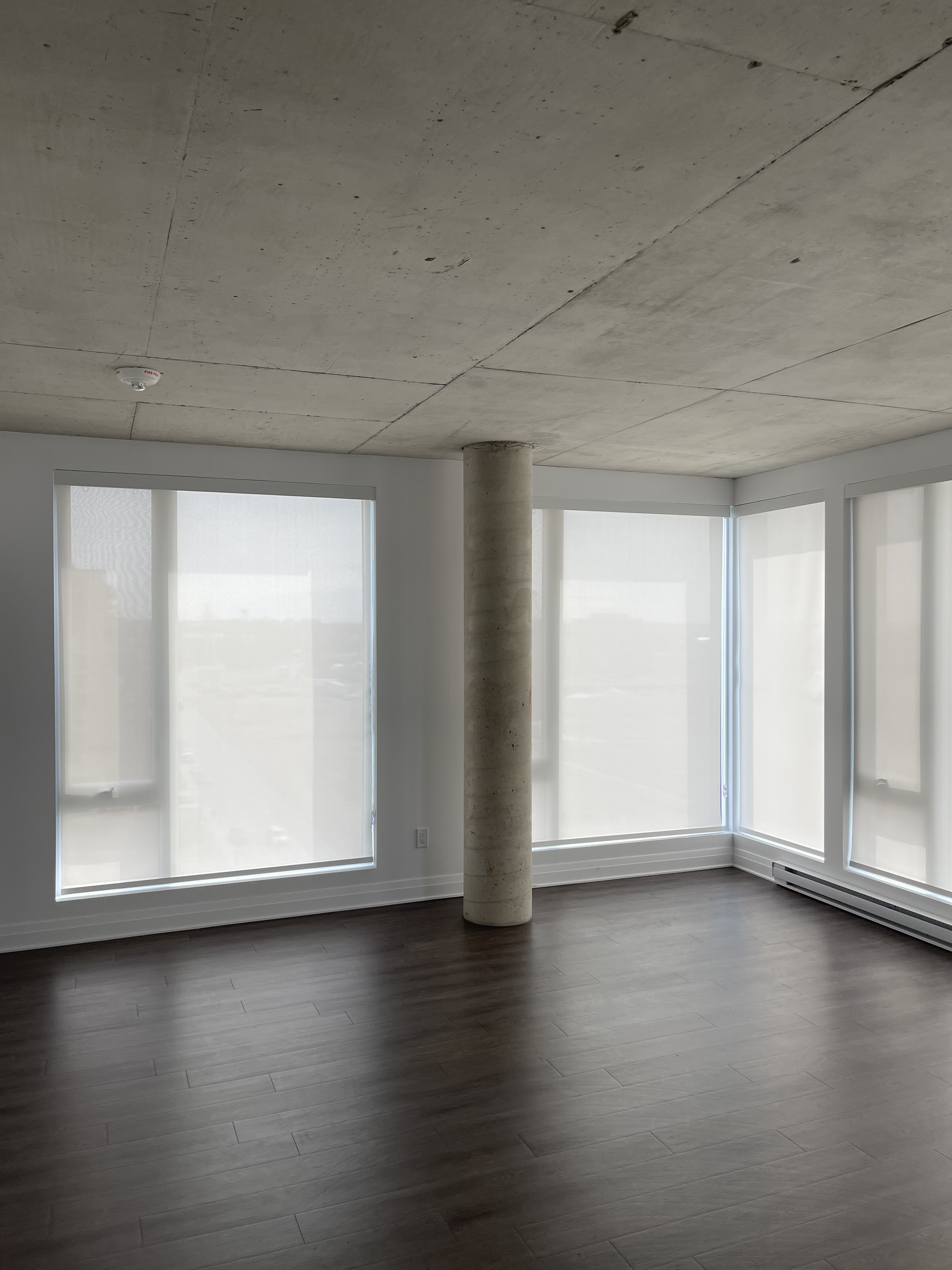 Empty modern apartment room with large windows covered by white blinds, dark wood flooring, a concrete ceiling, white walls, and a single cylindrical concrete pillar in the center.