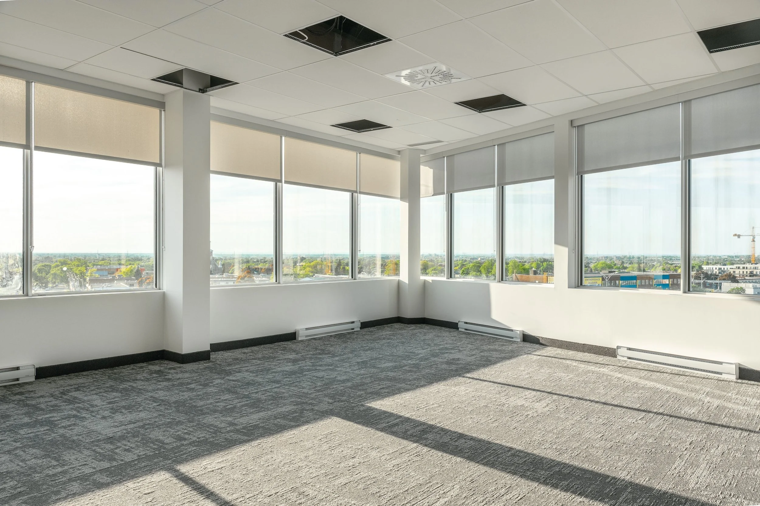 Empty office room with large windows and city view, some ceiling tiles missing, carpeted floor, and baseboard heaters.