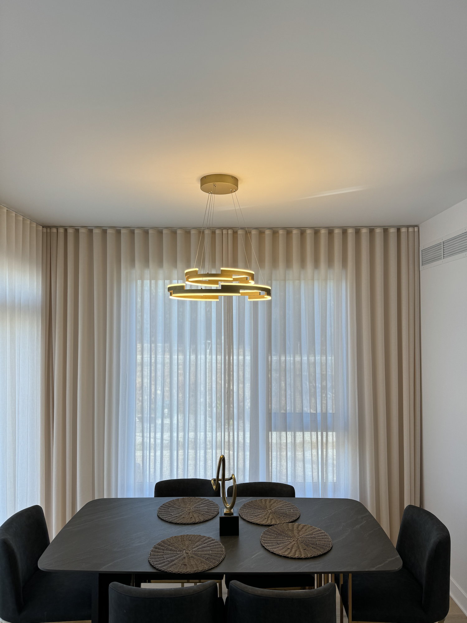 Modern dining room with black table, six black chairs, beige curtains, window, and contemporary gold chandelier.