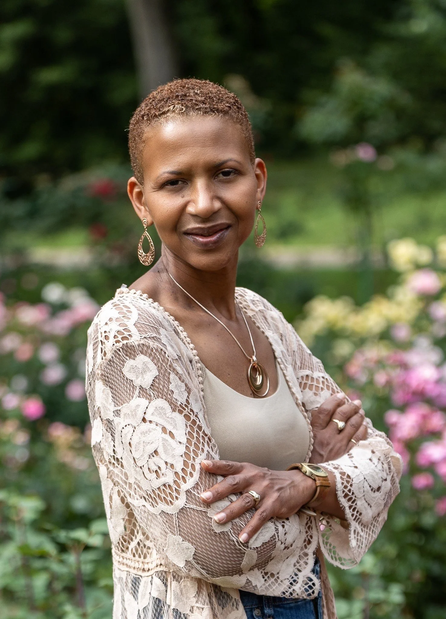Portrait of a woman with short, curly hair standing outdoors in a garden with pink flowers, wearing a lace jacket, a beige top, and jewelry including earrings, a necklace, and a watch.