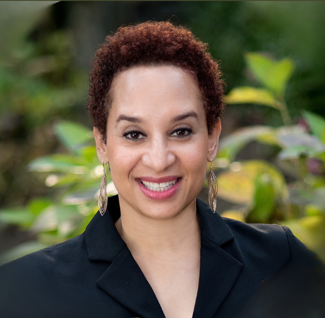 Close-up portrait of a woman with short curly hair, smiling, outdoors with greenery in the background.