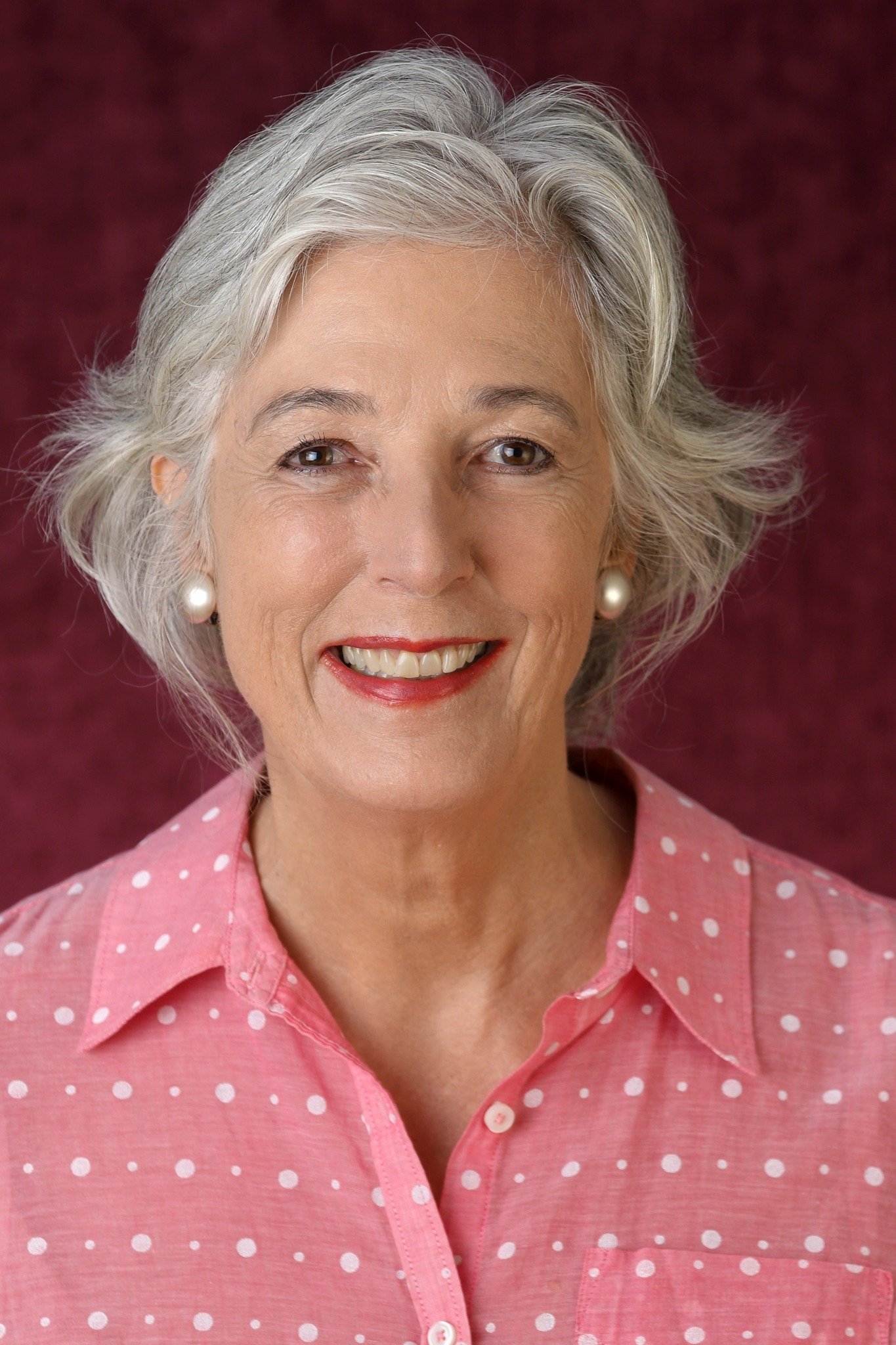 A smiling elderly woman with gray hair wearing pearl earrings and a pink polka dot shirt.