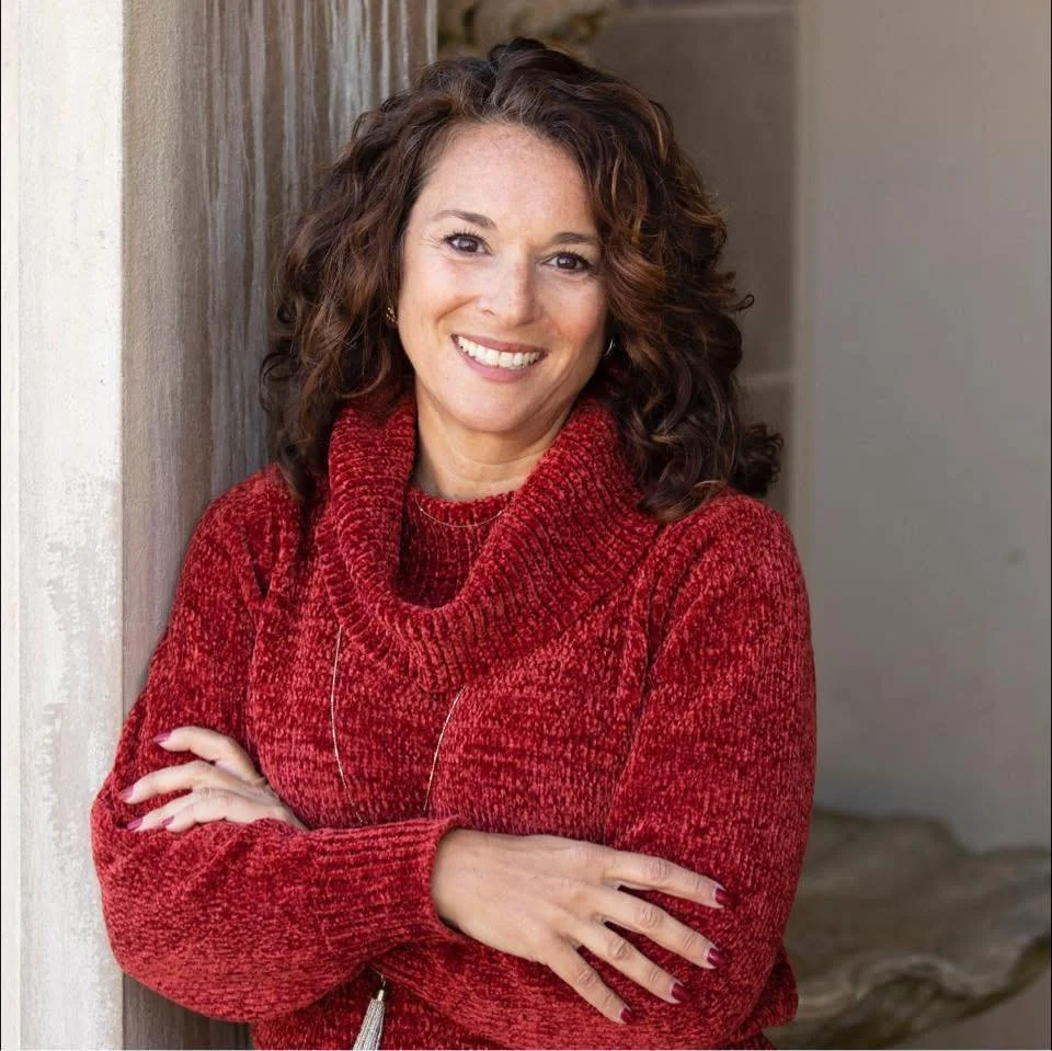 A woman with curly brown hair smiling and wearing a red knit turtleneck sweater, leaning against a wall.