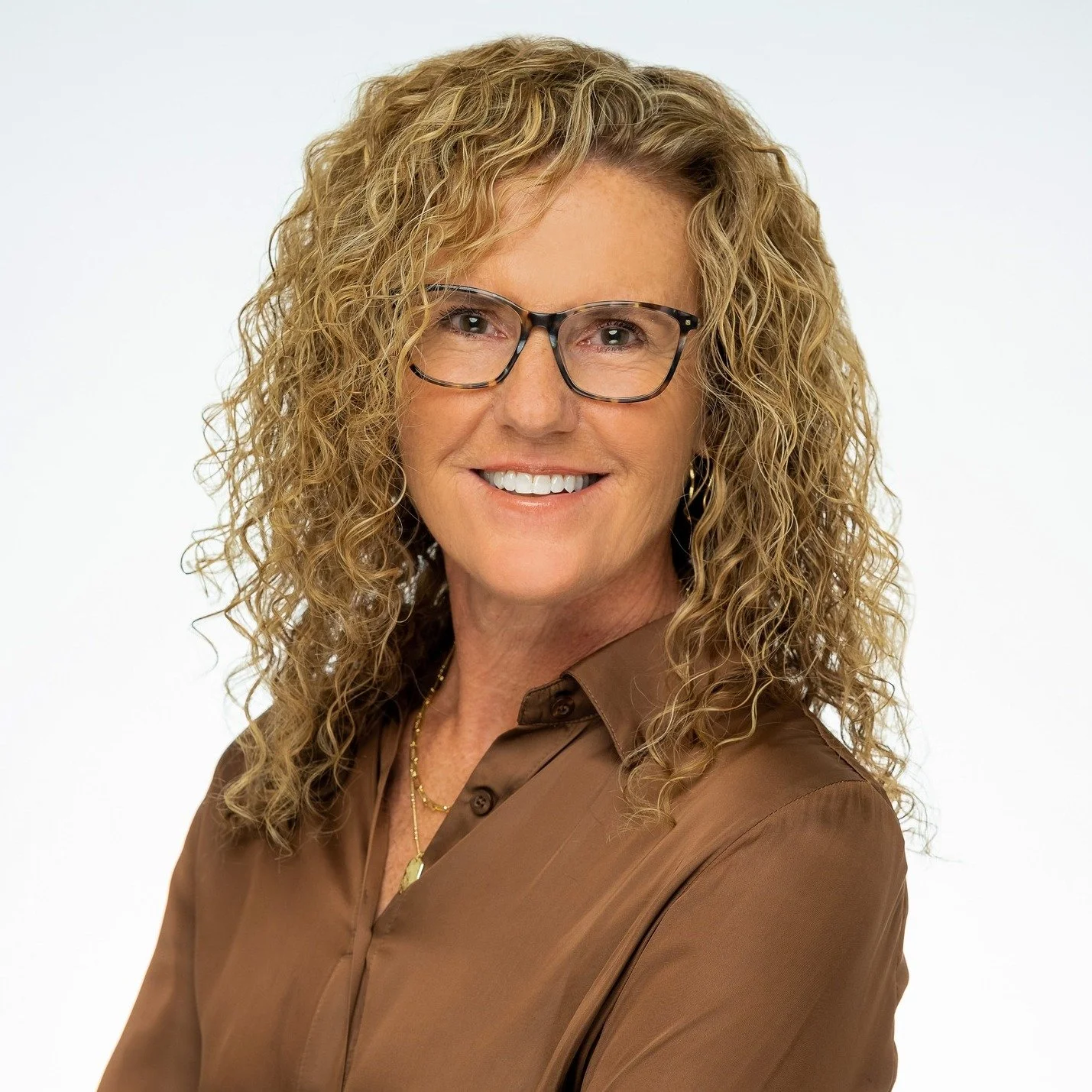 A woman with blond curly hair wearing glasses and a brown blouse, smiling against a white background.