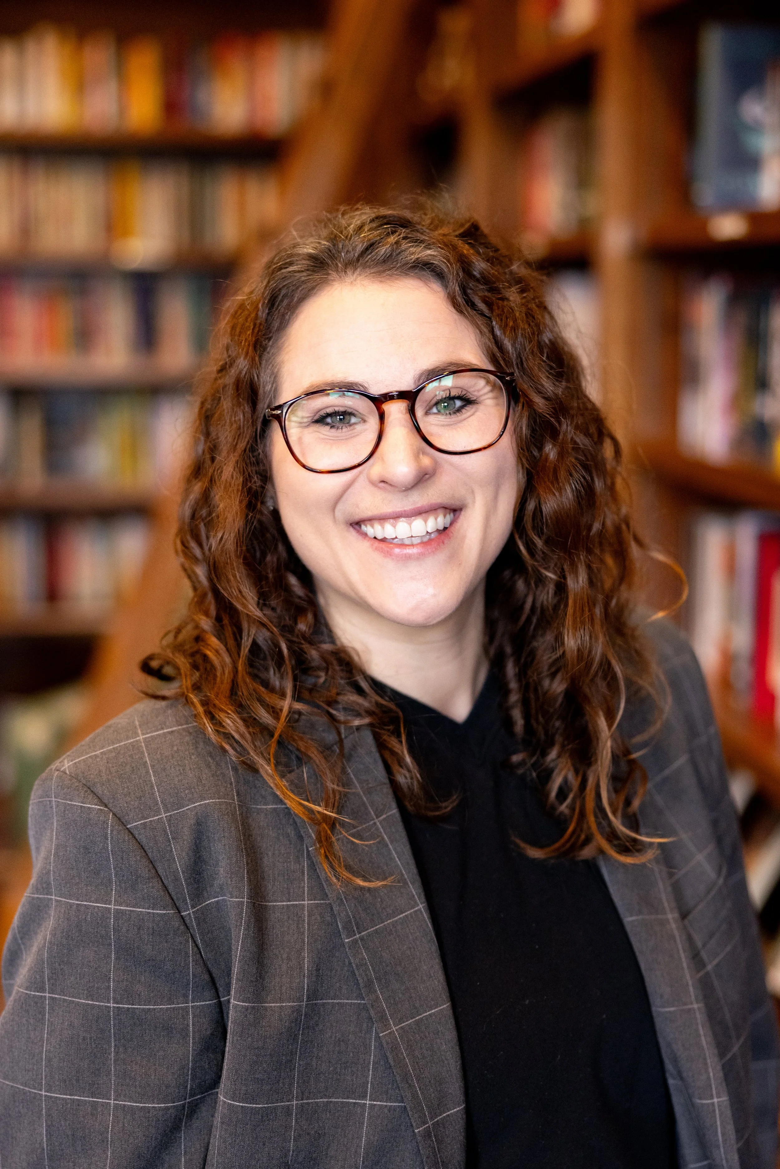 A woman with curly brown hair and glasses smiling in front of a library shelf filled with books.