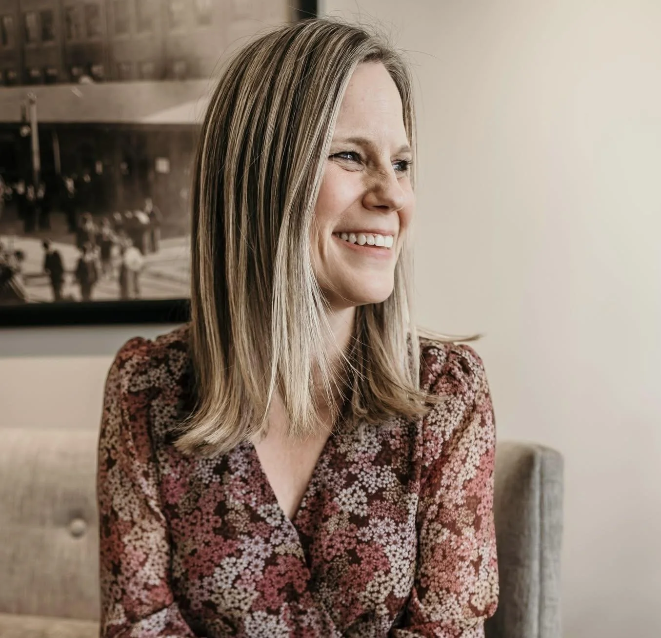 A smiling woman with shoulder-length blonde hair, wearing a floral patterned shirt, sitting on a sofa in a cozy room with a framed black-and-white photo in the background.