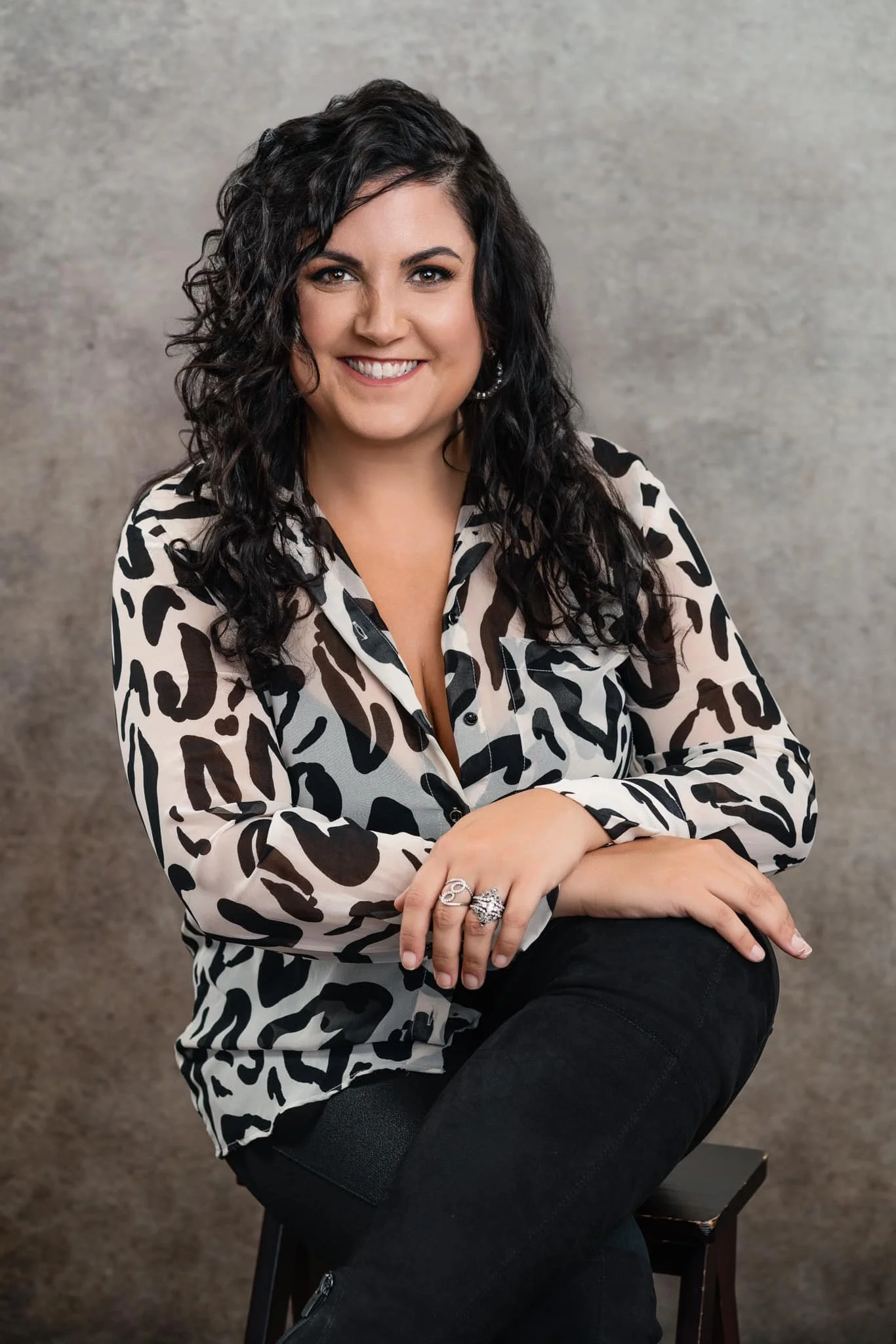 A woman with curly black hair, wearing a black and white animal print blouse, sitting on a stool against a neutral background, smiling at the camera.